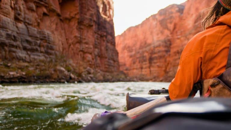 A rafter in an orange jacket paddles through choppy water between towering red canyon walls.