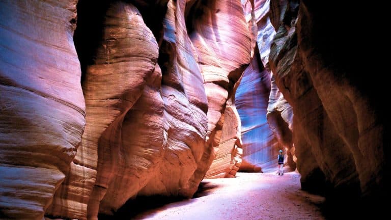 A lone hiker walks through a narrow slot canyon with towering, colorful sandstone walls glowing in shades of pink, red, and purple.