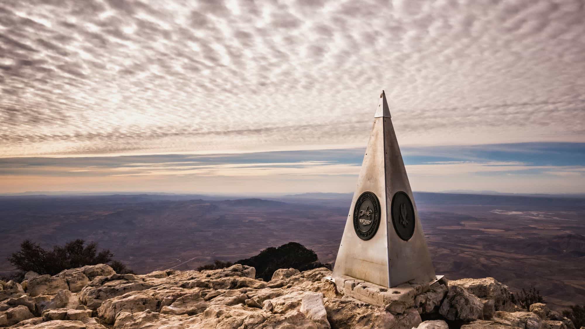 A silver pyramid monument rises on rocky ground at the summit of Guadalupe Peak, overlooking a sweeping desert landscape.