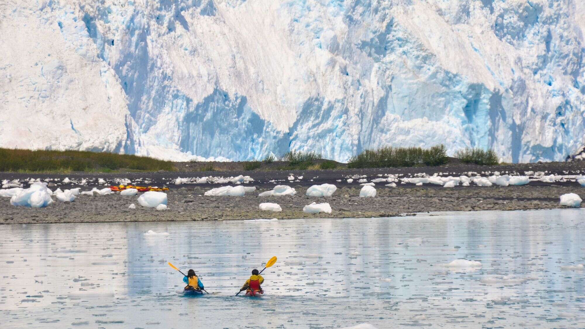 Two kayakers paddle through icy waters with scattered ice chunks in front of a massive glacier.