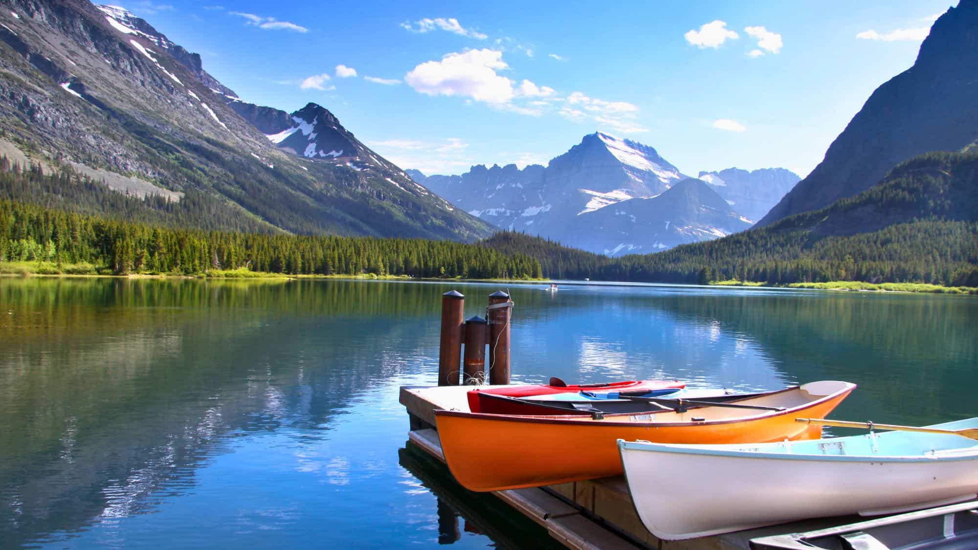 Colorful canoes rest on a dock overlooking a serene lake surrounded by snow-capped peaks and dense forest.