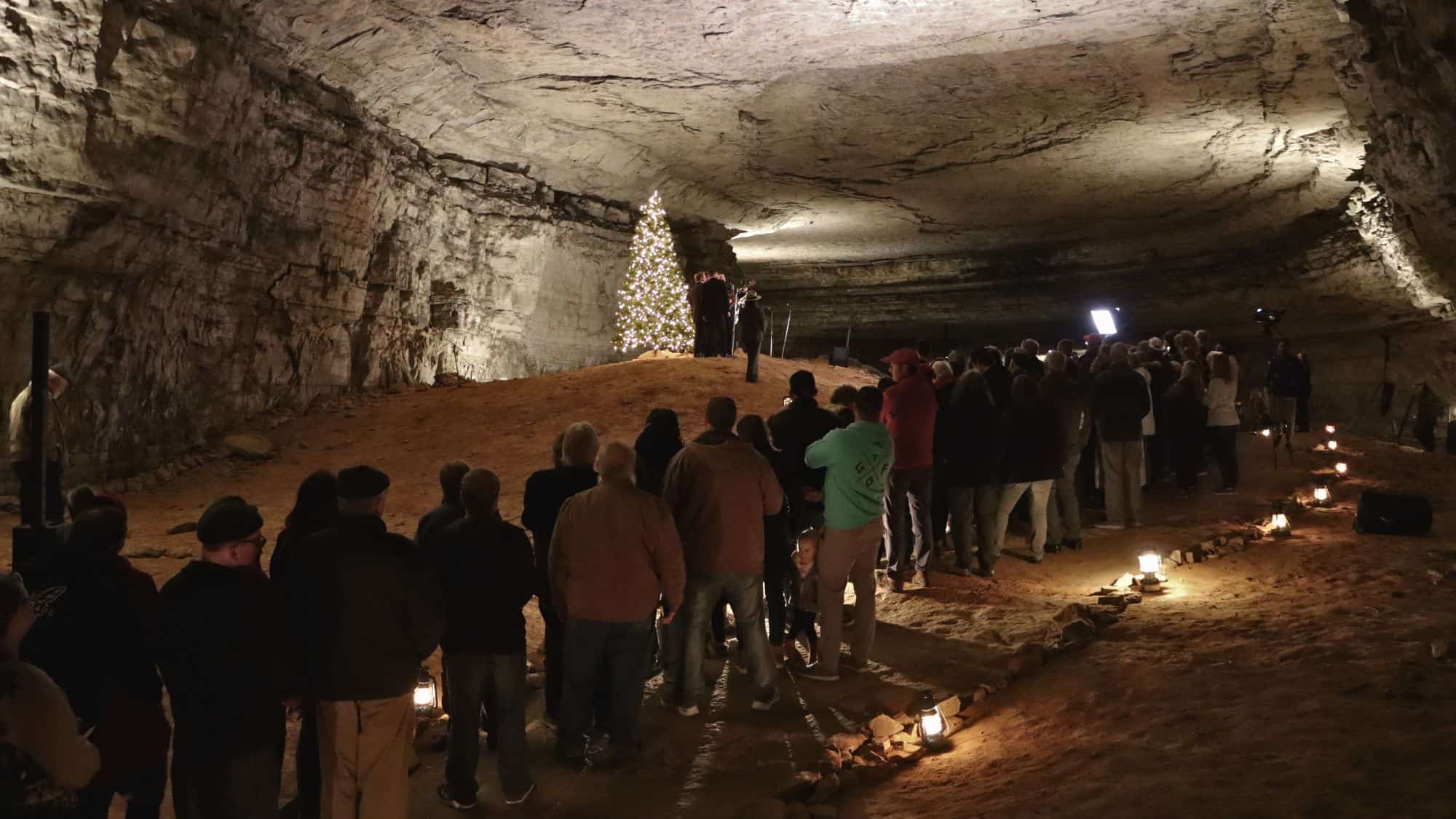 A crowd gathers inside a cavern decorated with a lit Christmas tree, watching a small group perform on stage.