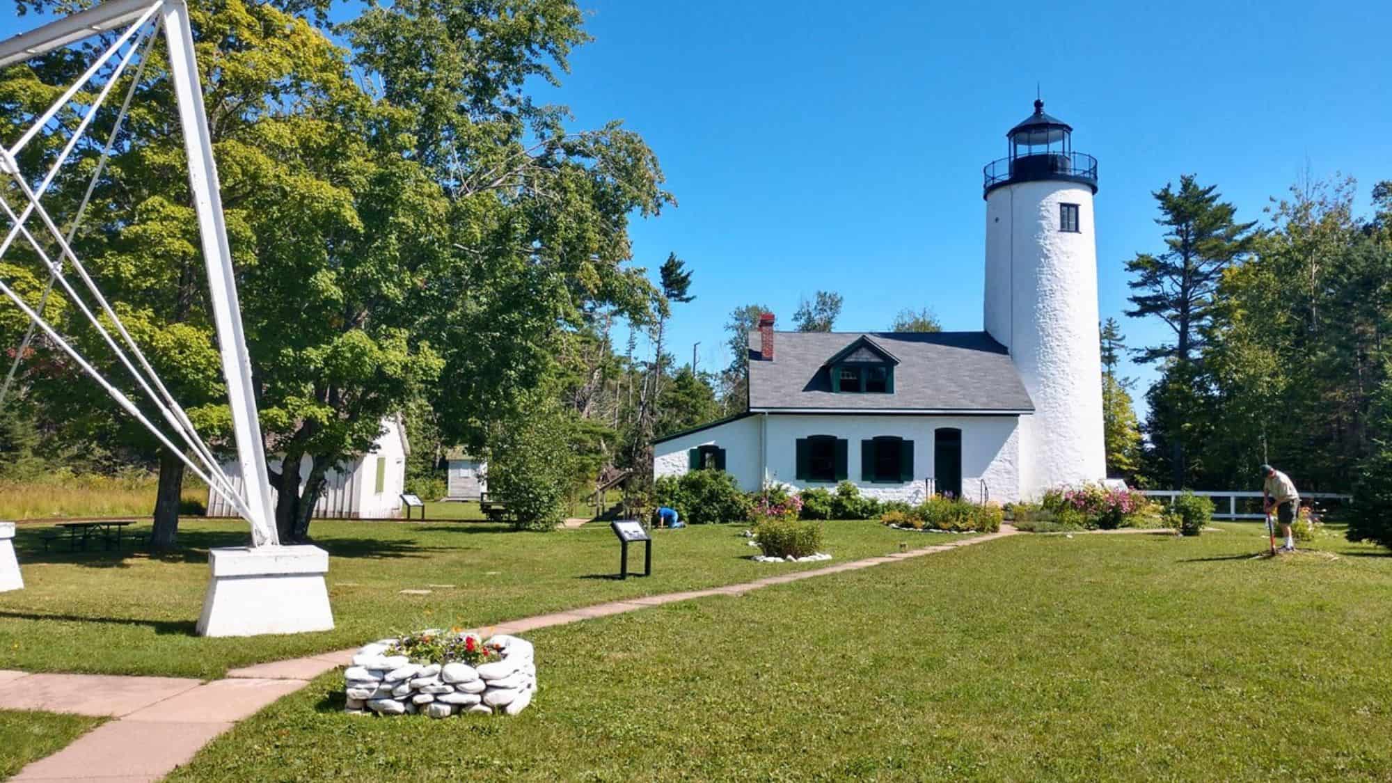 A round white lighthouse and cottage sit in a grassy garden, surrounded by trees and flowerbeds on a bright, sunny day.