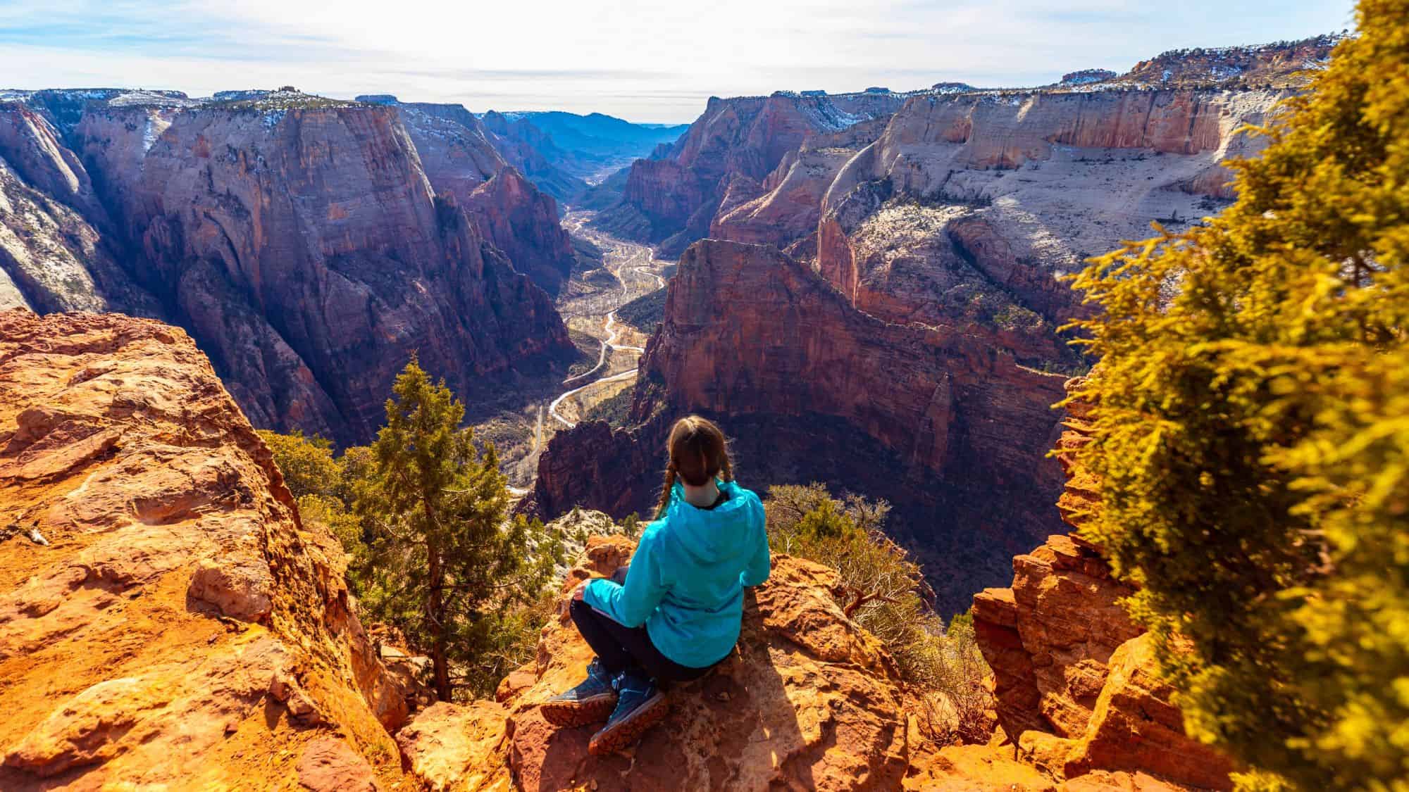 A hiker in a blue jacket sits on a cliff edge, looking out over the towering red rock walls and winding valley of Zion National Park.