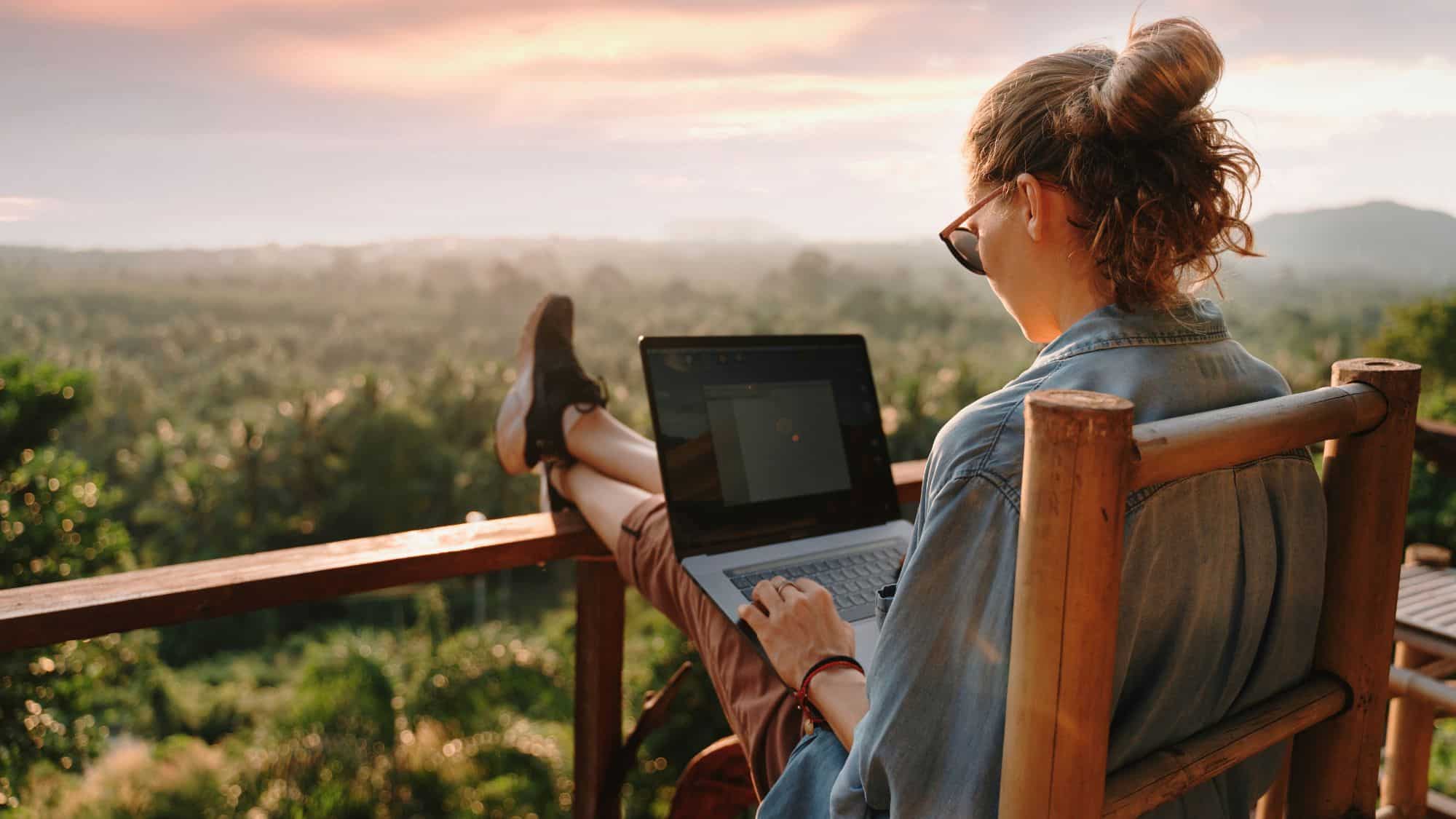 A woman reclines on a wooden chair with her laptop, gazing out at a lush green landscape under a glowing sunset sky.