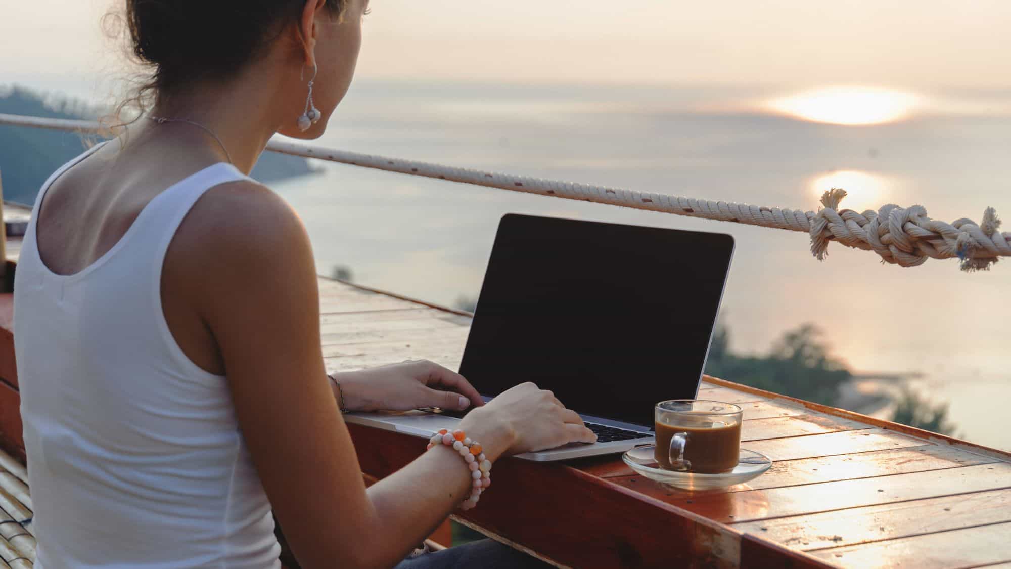 A woman types on her laptop at a wooden counter with a cup of coffee nearby, overlooking the ocean at sunset.