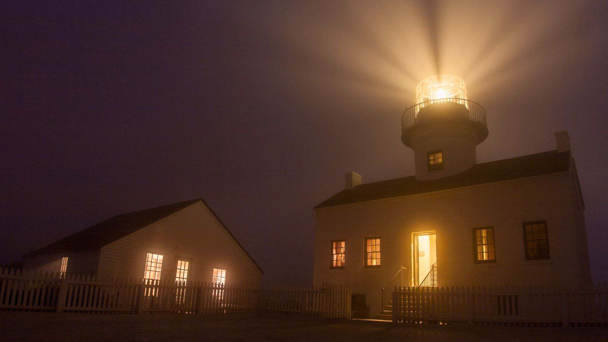 A lighthouse shines brightly through thick fog at night, its beam casting long rays across the dark sky above a white picket fence.