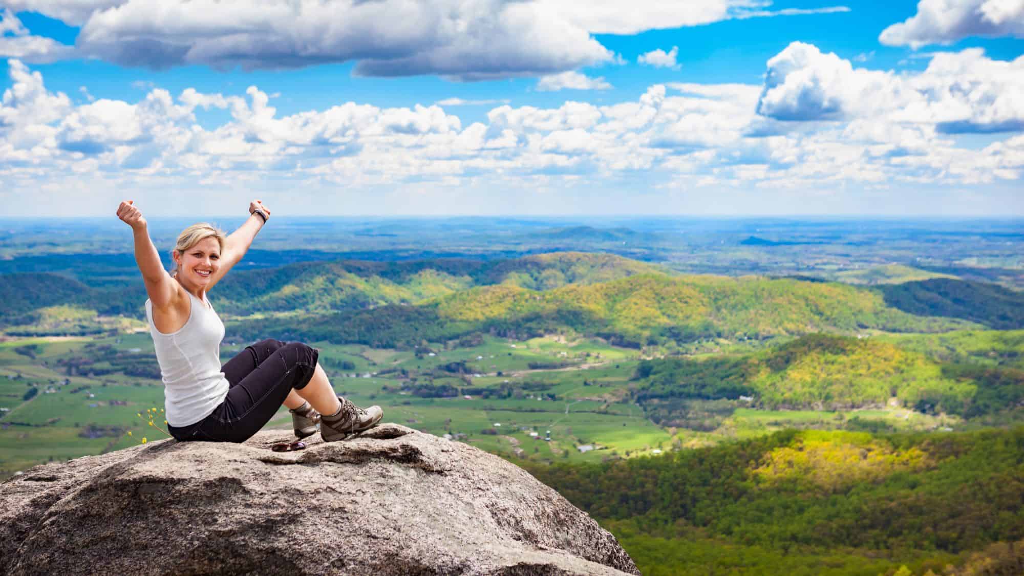 A smiling woman in hiking gear raises her arms in triumph while sitting on a boulder overlooking a vast green valley.