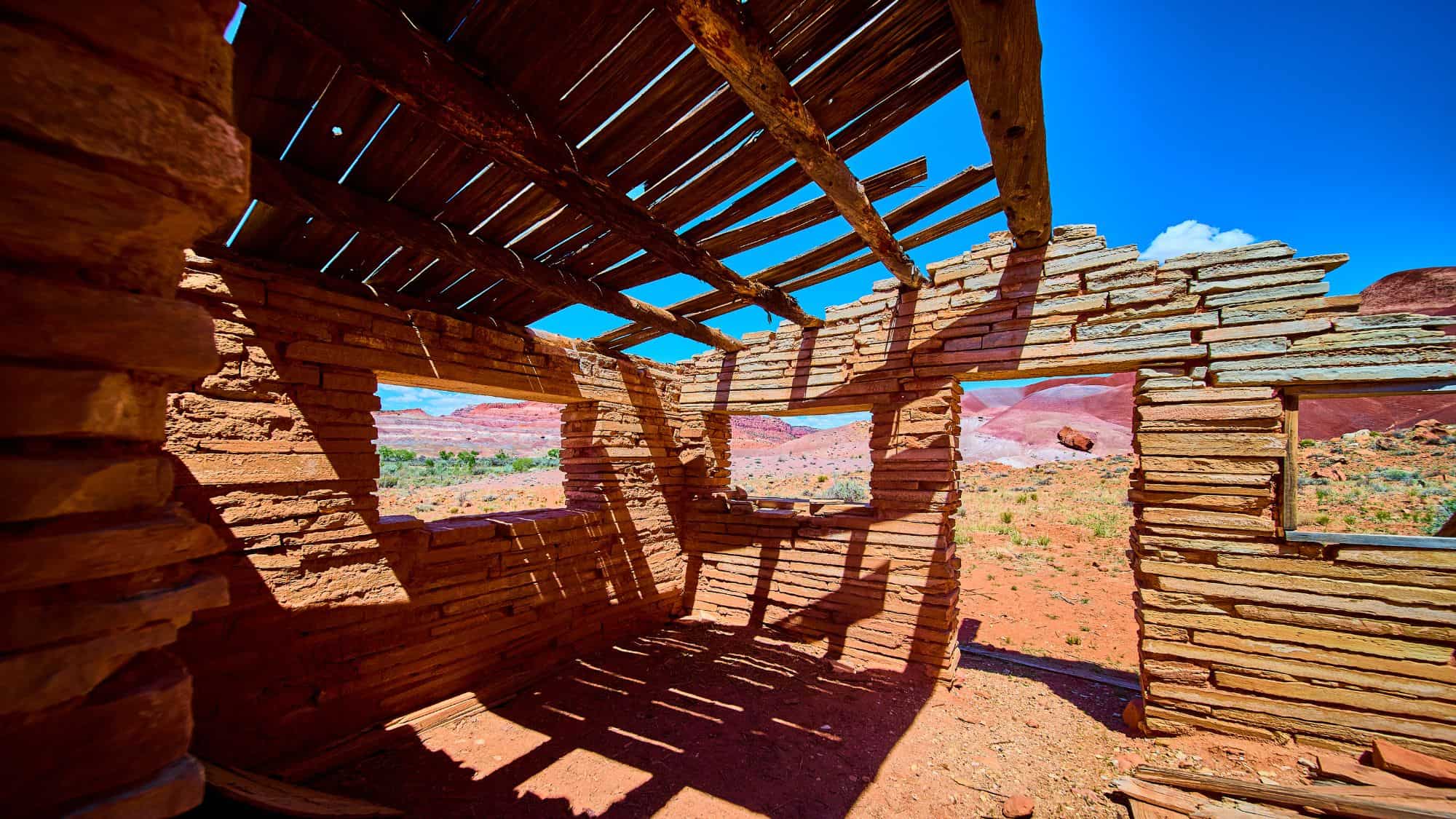 The sun casts long shadows through the remains of a sandstone cabin with a collapsed roof, framing a view of vivid red desert hills beyond.