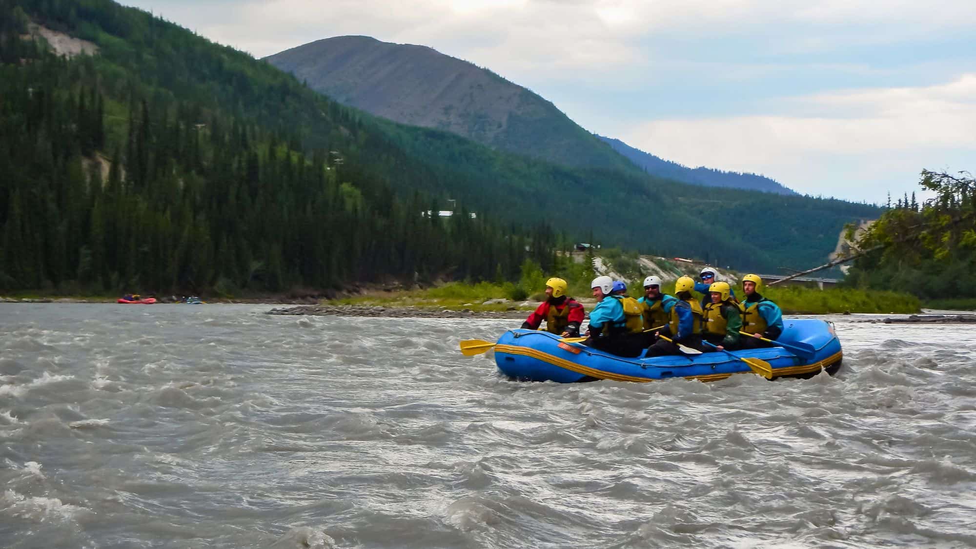 A group of rafters in helmets and life jackets navigates rough waters against a backdrop of forested mountains.