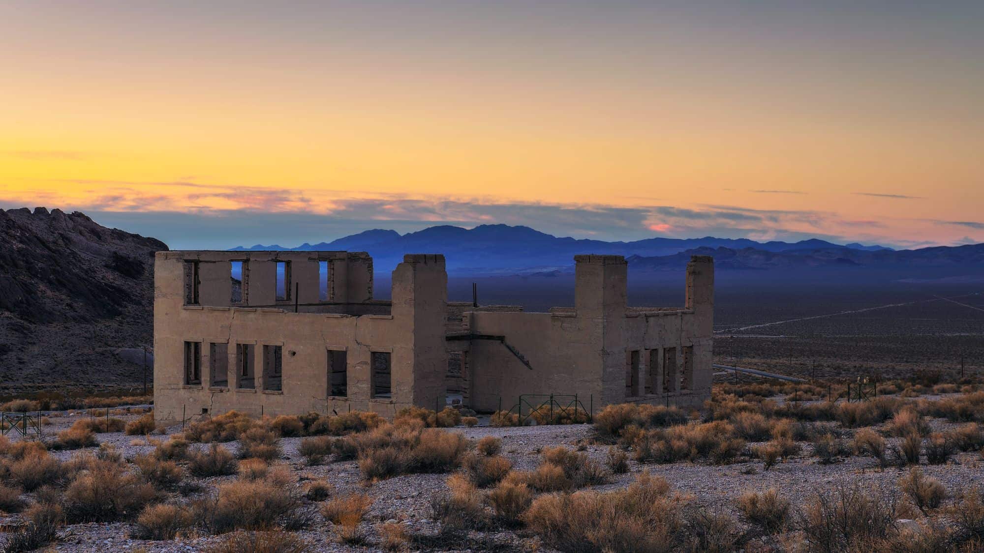 Crumbling remains of a concrete building stand alone in the Nevada desert at twilight, with distant purple mountains silhouetted under a fading sunset sky.