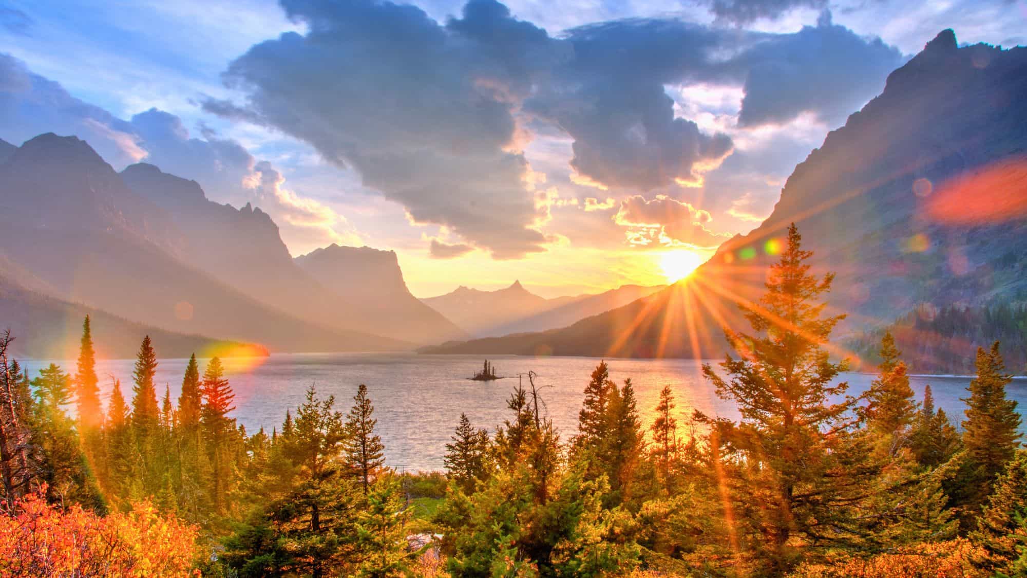 The sun bursts over a distant mountain ridge, casting golden light across a tranquil lake surrounded by evergreen trees and dramatic peaks.