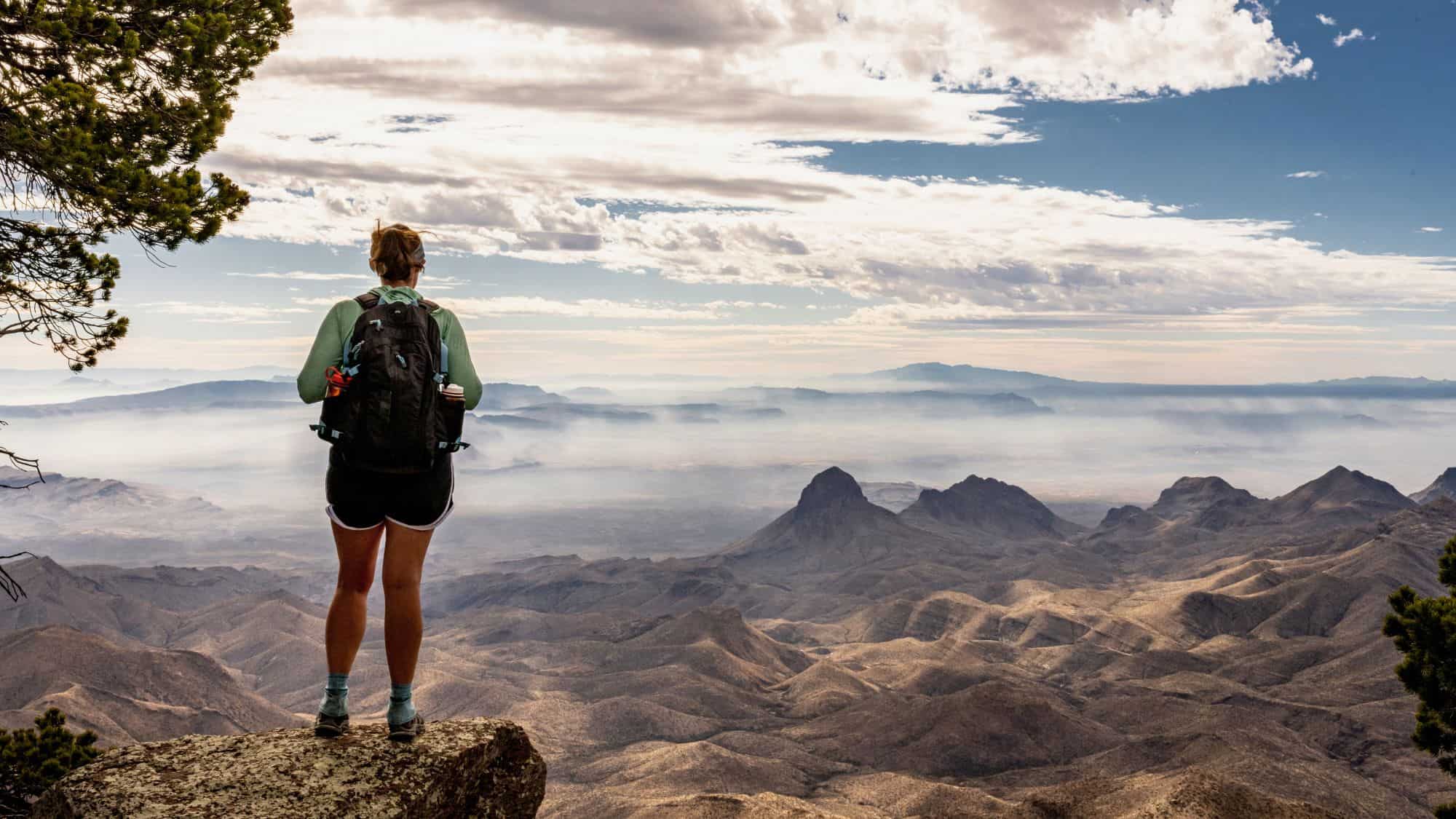 A hiker with a backpack stands on a rocky ledge gazing out at a sweeping view of hazy desert peaks beneath a cloud-filled sky.