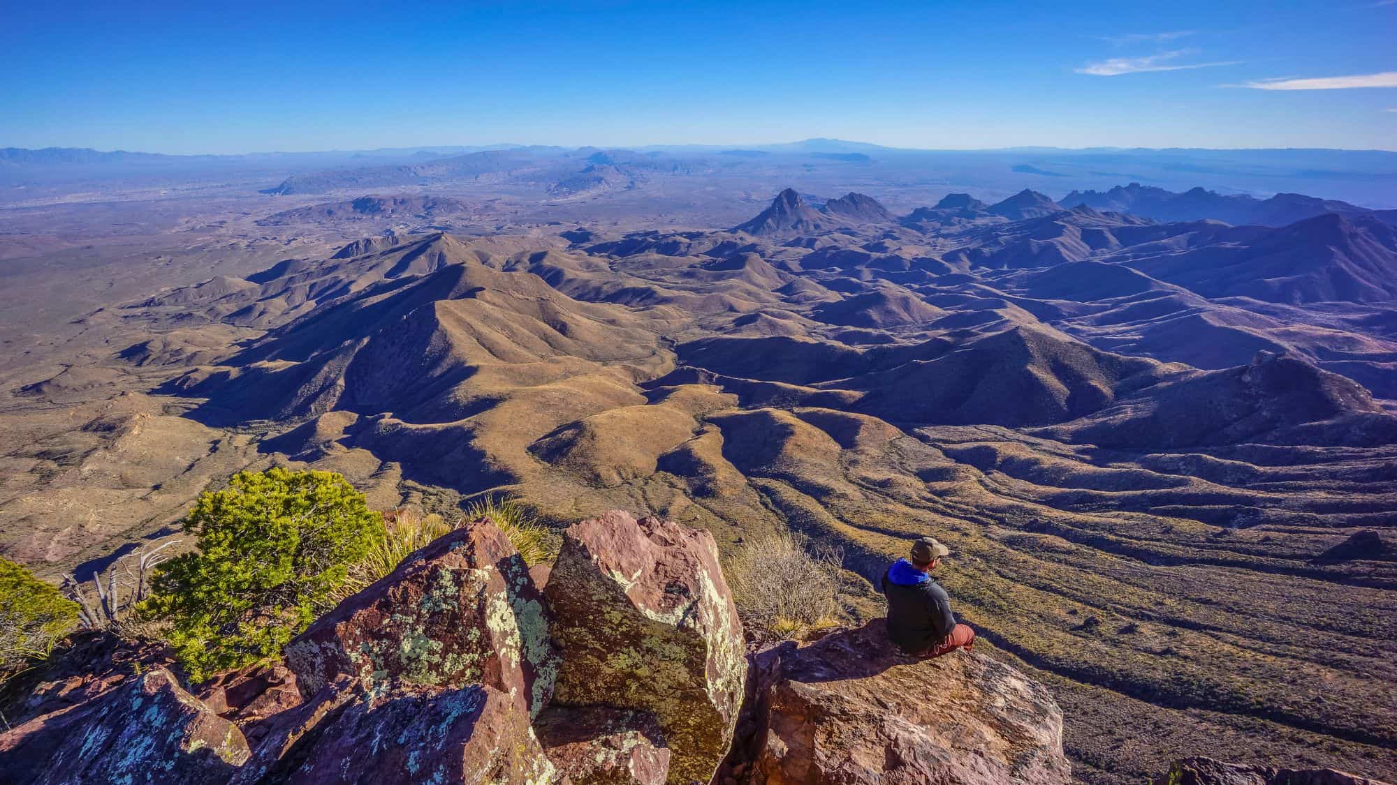 A lone hiker perches on rocks high above Big Bend’s rugged mountain ridges, stretching endlessly across the desert.
