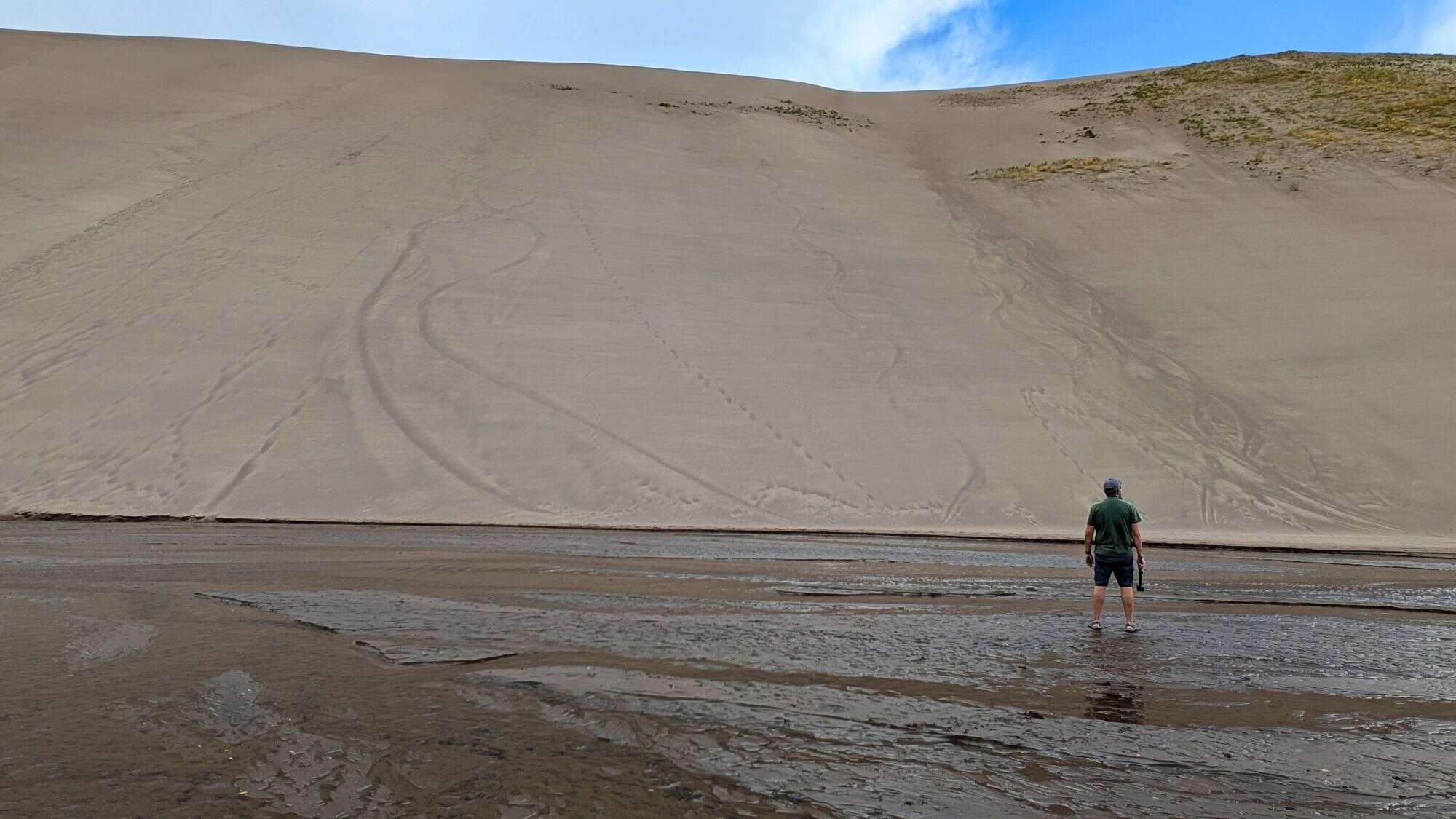 A lone person stands at the base of a towering sand dune with rippling patterns etched into its slope.