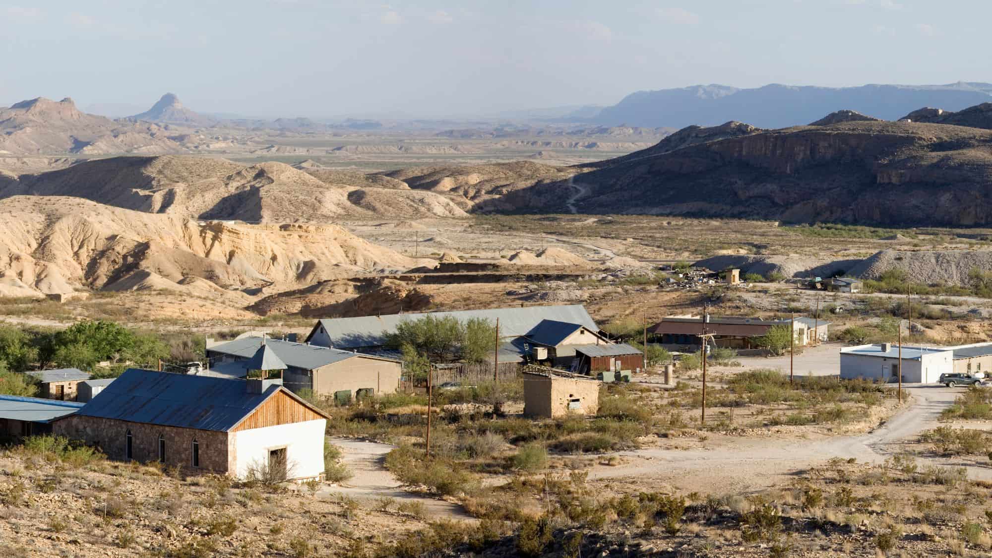 A scattering of adobe and metal-roofed buildings lie across a dusty landscape in Terlingua, Texas, with pale desert hills stretching to the horizon.