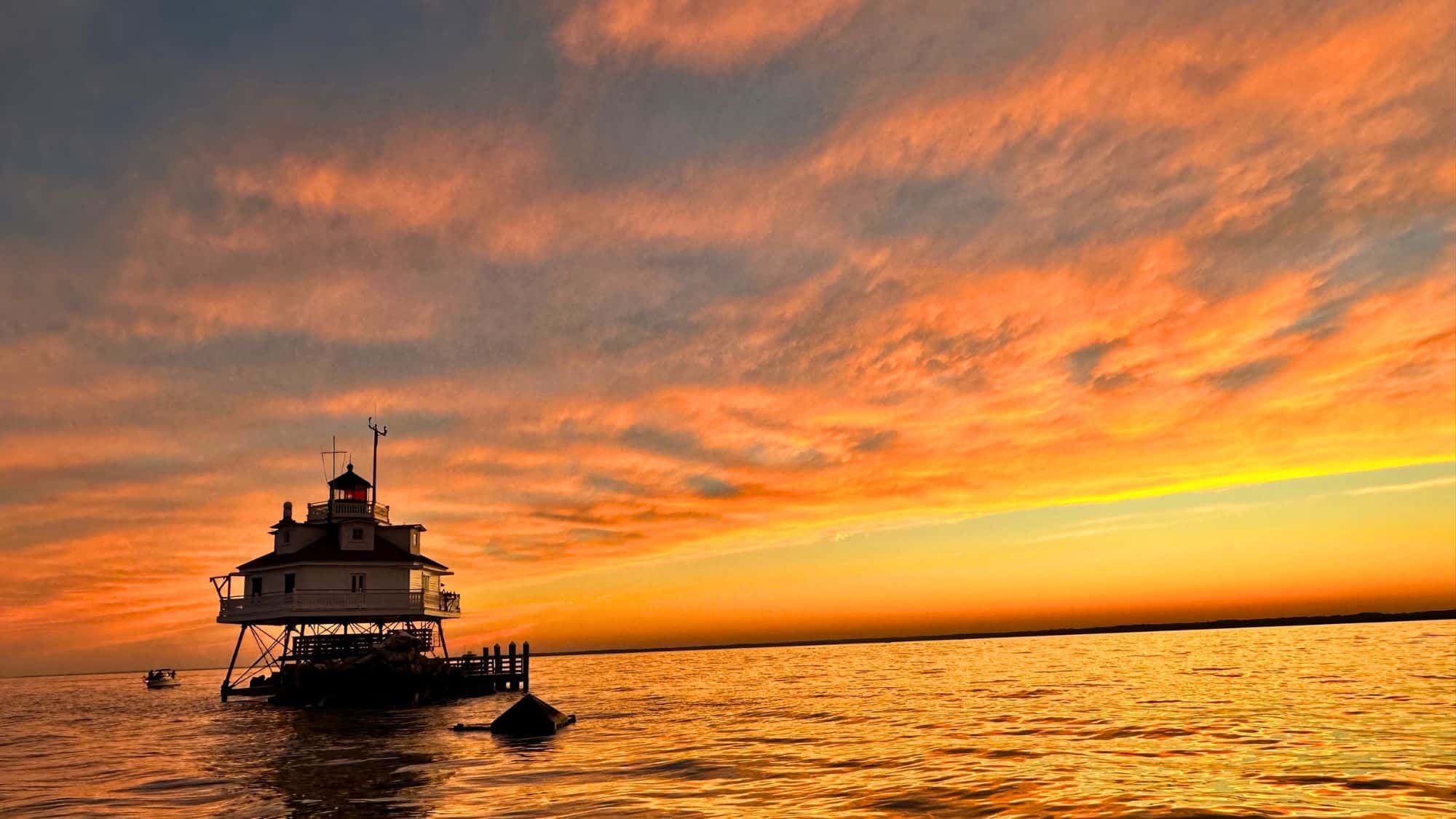 A screw-pile lighthouse on stilts is silhouetted against a vibrant orange and pink sunset over calm waters in the Chesapeake Bay.