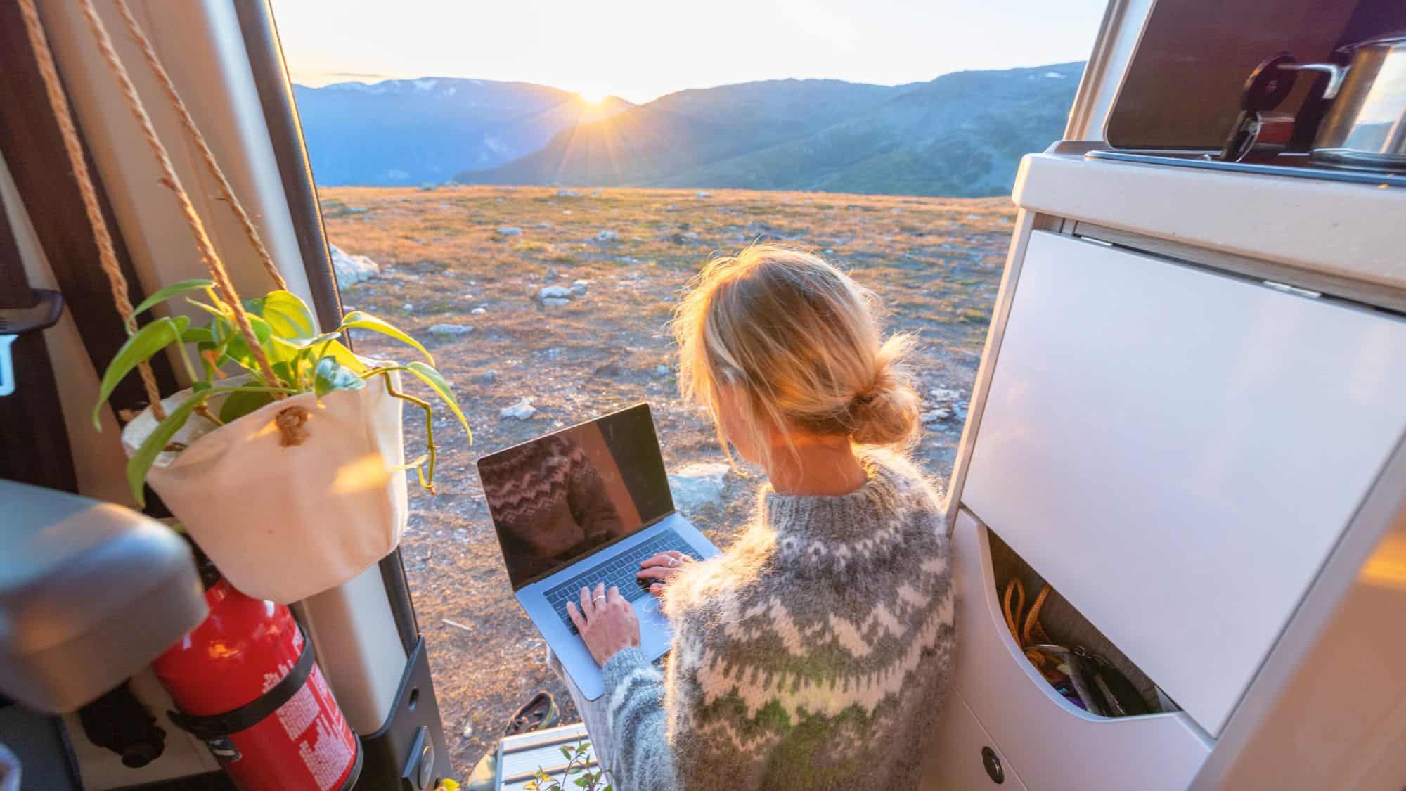A woman in a patterned sweater works on her laptop from the open door of a camper van with mountains and a rising sun in the background.
