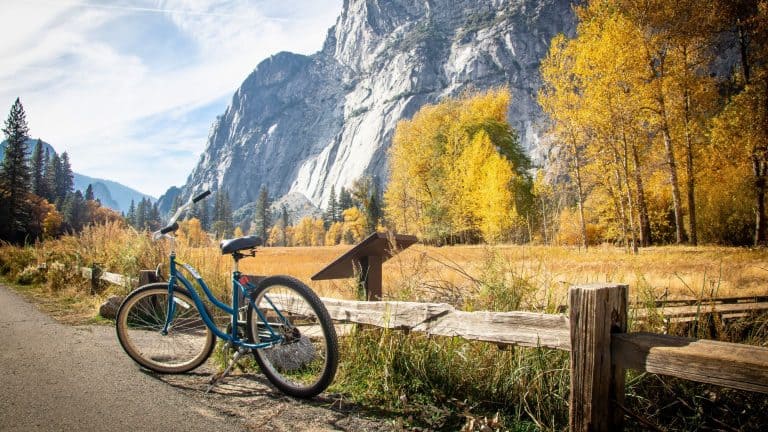 A blue bicycle rests against a wooden fence with golden autumn trees and dramatic granite cliffs in the distance.