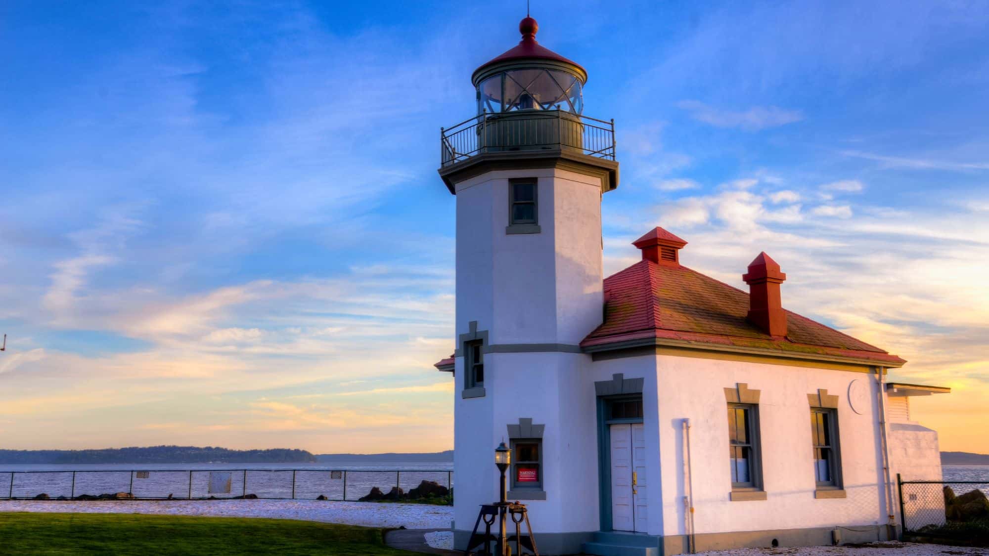 A small white lighthouse with a red roof sits beside Puget Sound, glowing in soft evening light beneath a pastel sky.