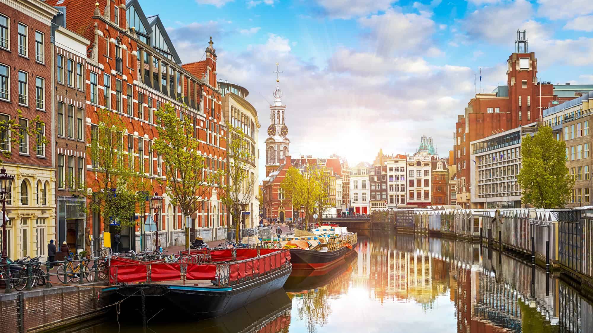 Boats line a sunlit canal in Amsterdam, bordered by historic Dutch buildings and a church spire.