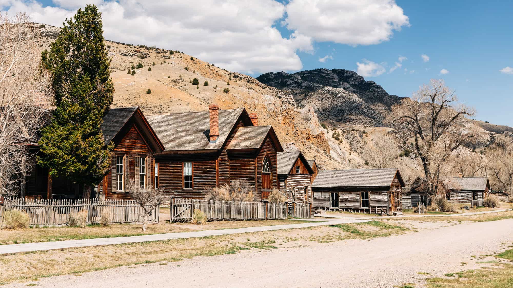 A row of well-preserved wooden buildings with picket fences lines a dirt road in Bannack, Montana, surrounded by dry hills and rocky outcrops.