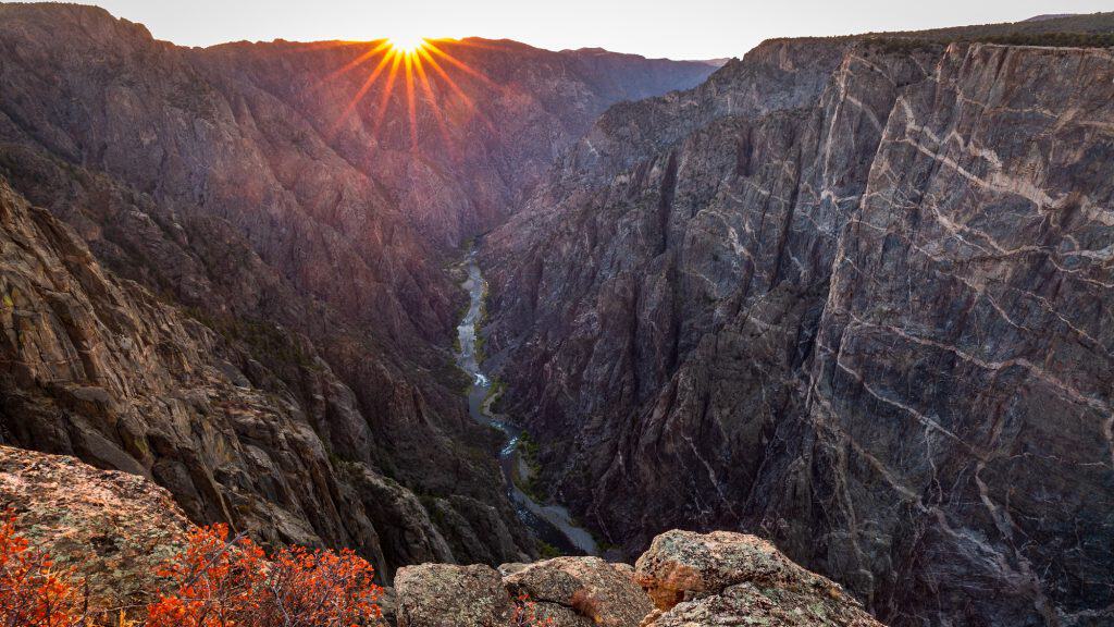 The sun sets over the steep cliffs of Black Canyon, casting golden light across rugged rock walls with a winding river flowing far below.