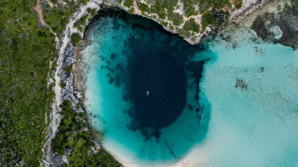A person floats in the center of a deep blue hole surrounded by turquoise shallows and lush greenery along the shore.