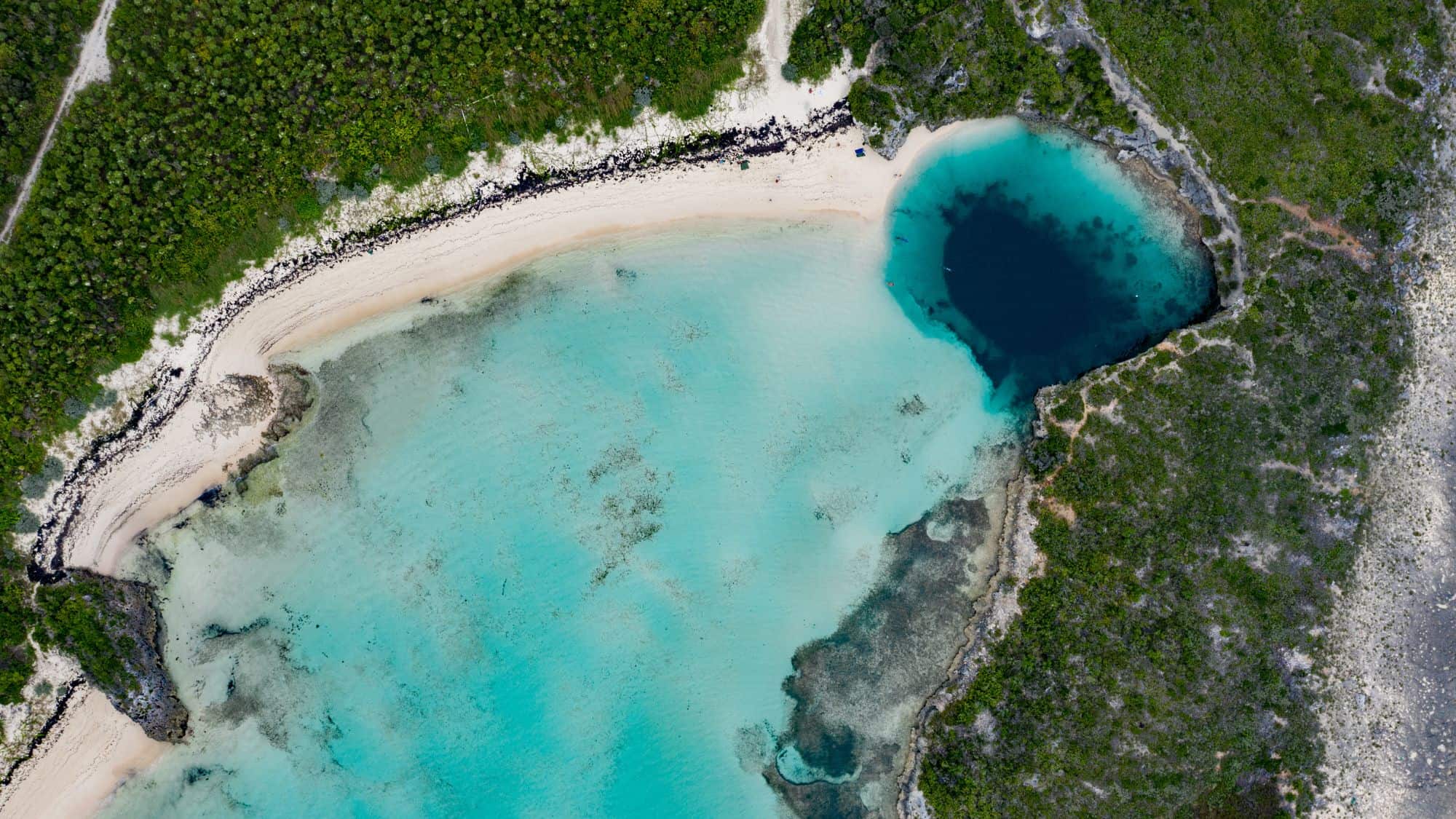 Aerial view of a tropical coastline with turquoise shallows and a deep circular blue hole near the beach.