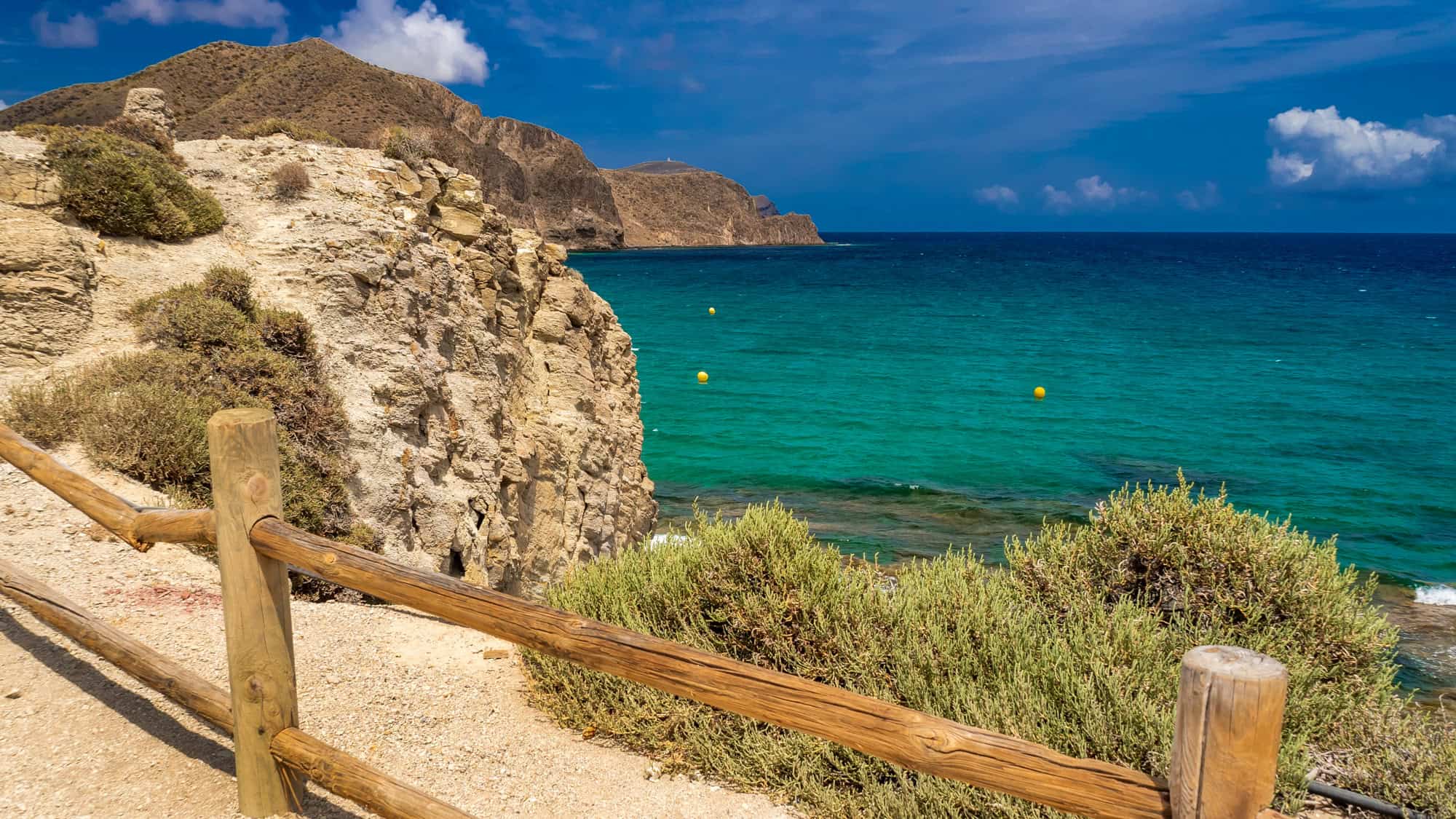 A wooden fence lines a sandy path along a rugged Mediterranean coastline, overlooking turquoise waters and rocky cliffs.