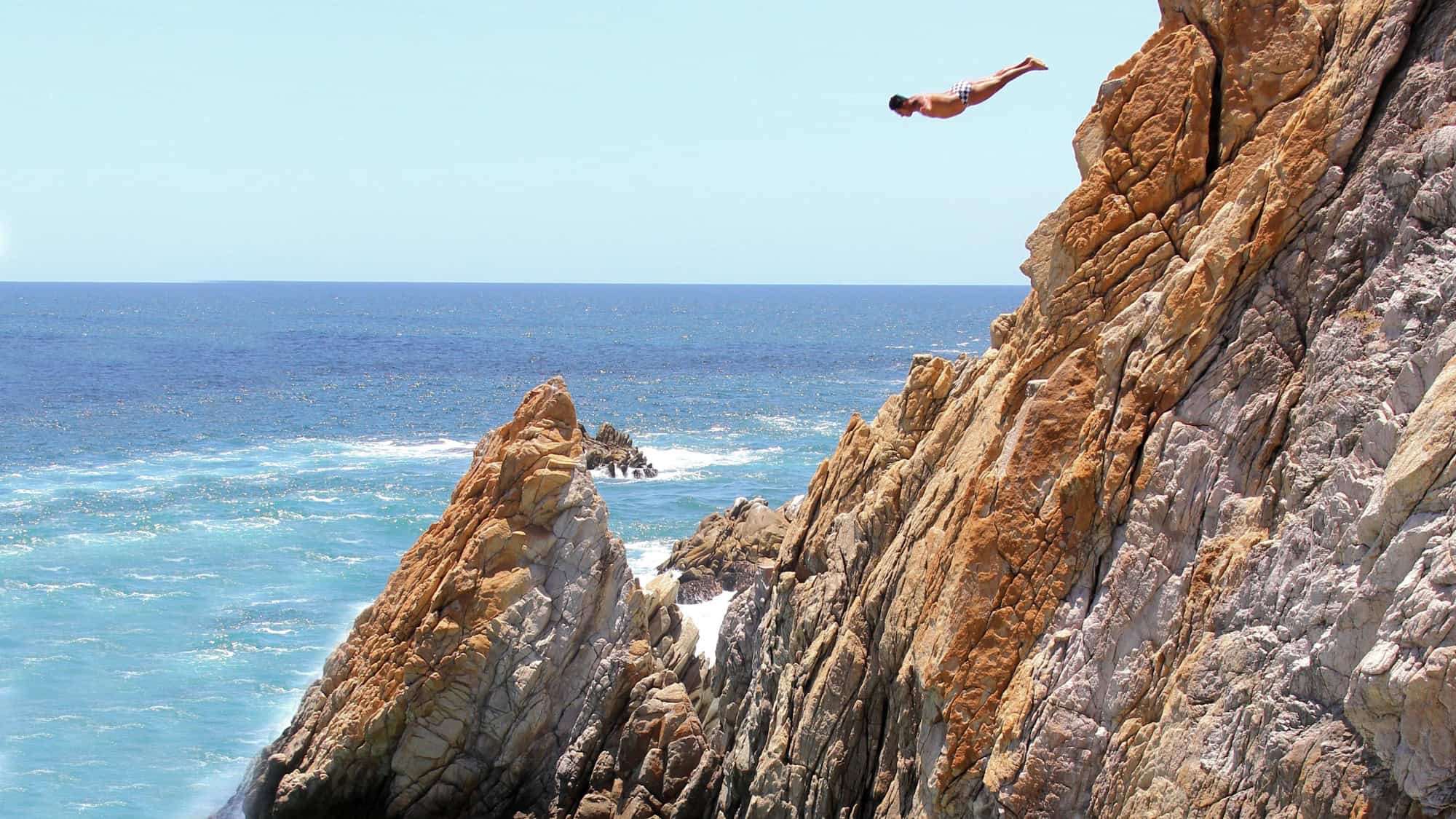 A man dives headfirst off a tall rocky cliff into the ocean below, with waves crashing against the rocks.