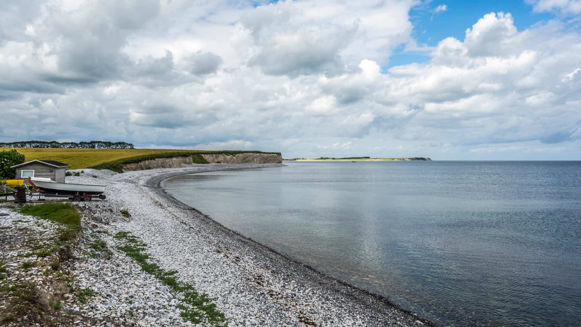 A small boat rests near a shoreline cabin beside a peaceful, curved pebble beach and calm, glassy waters under cloudy skies.