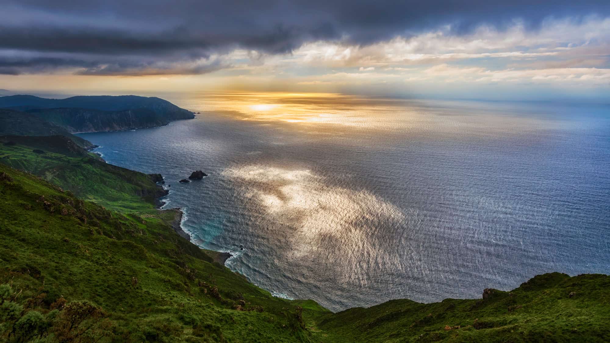 Golden light reflects on the ocean below a lush green headland, with dark clouds parting to reveal a glowing sky.