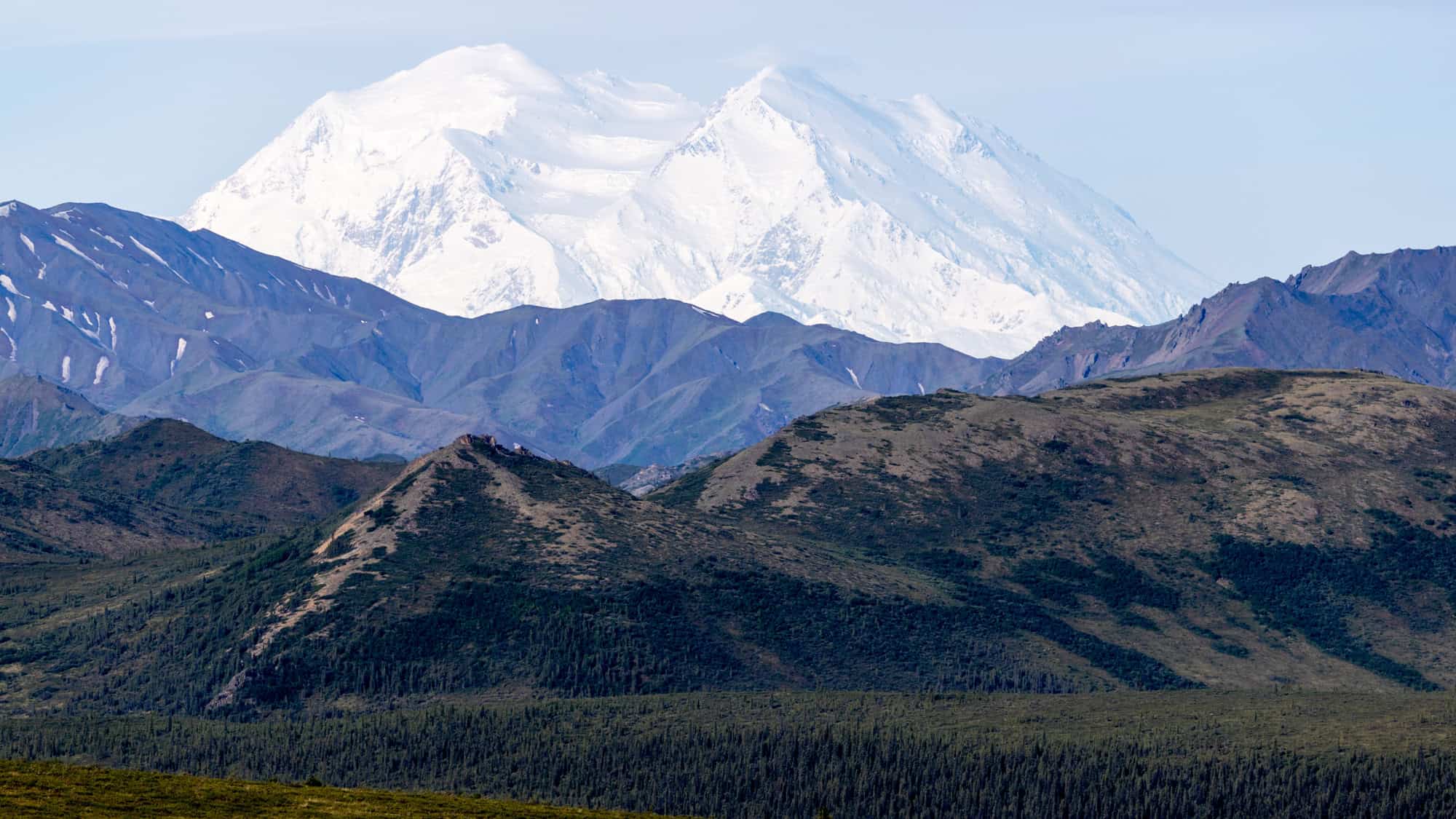 Towering white mountains rise behind dark, rugged foothills covered in forests and ridges.