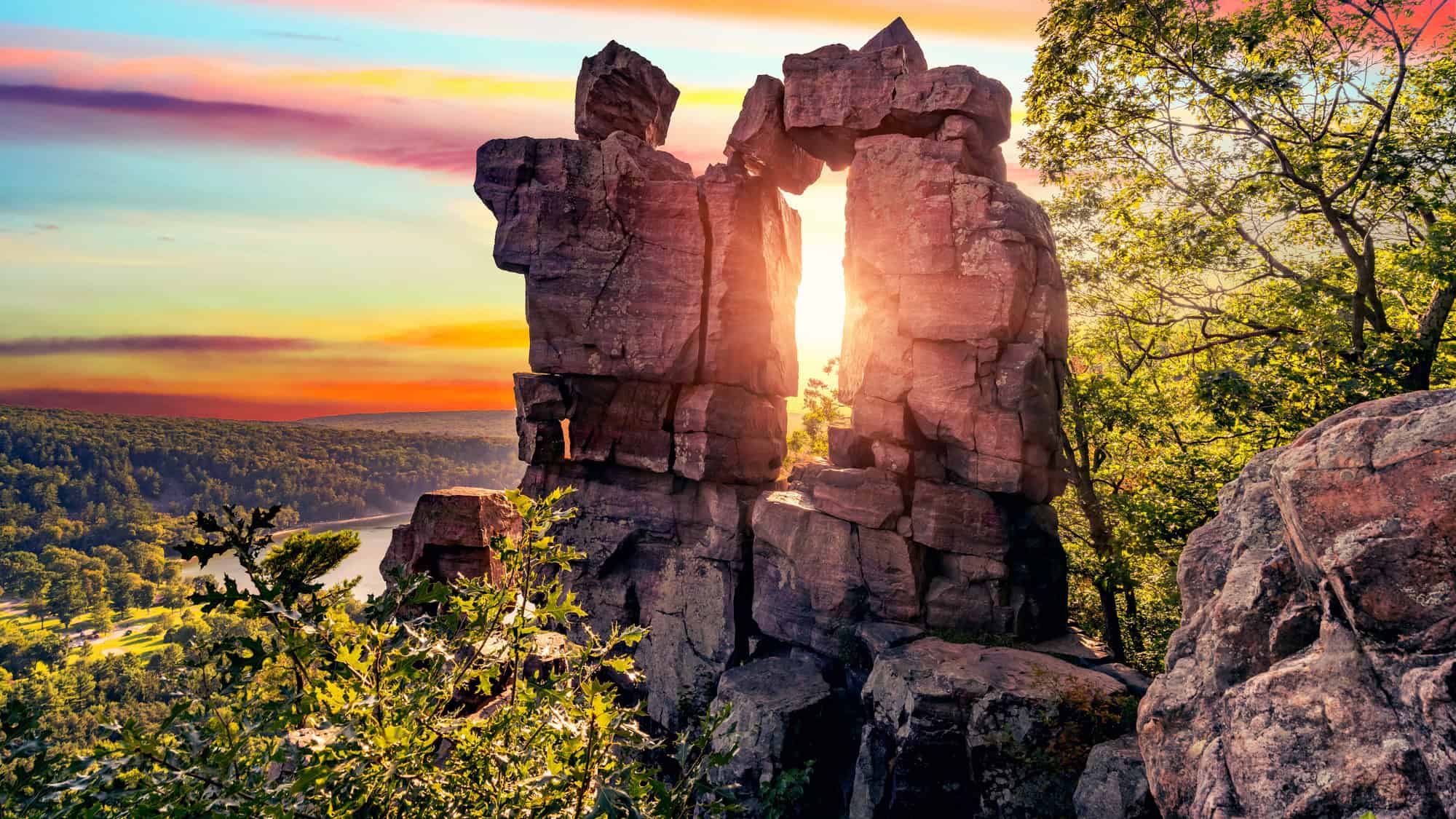 A dramatic rock formation frames the setting sun, with colorful skies and forested hills rolling into the distance.