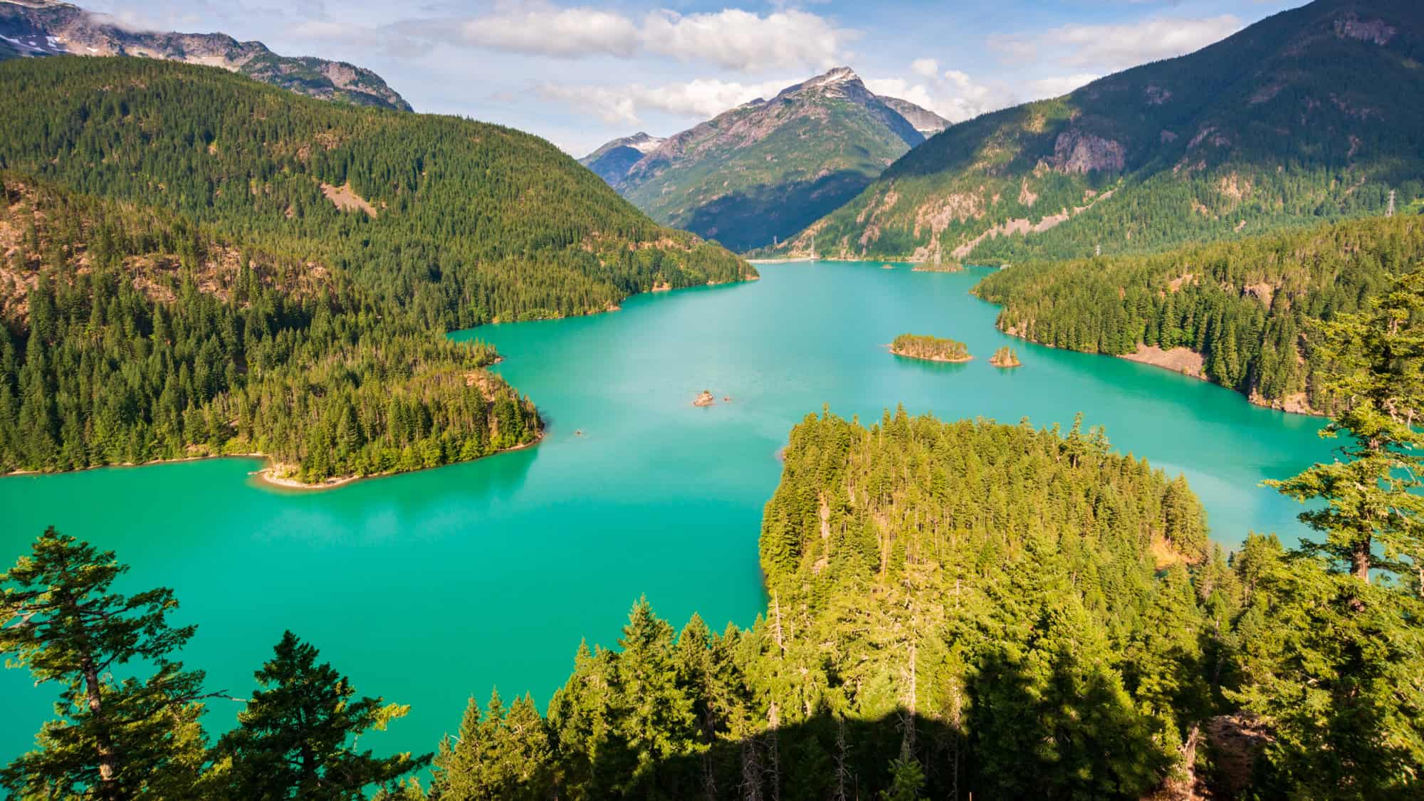 A vivid emerald-green lake winds between forested ridges and mountain peaks under scattered clouds.
