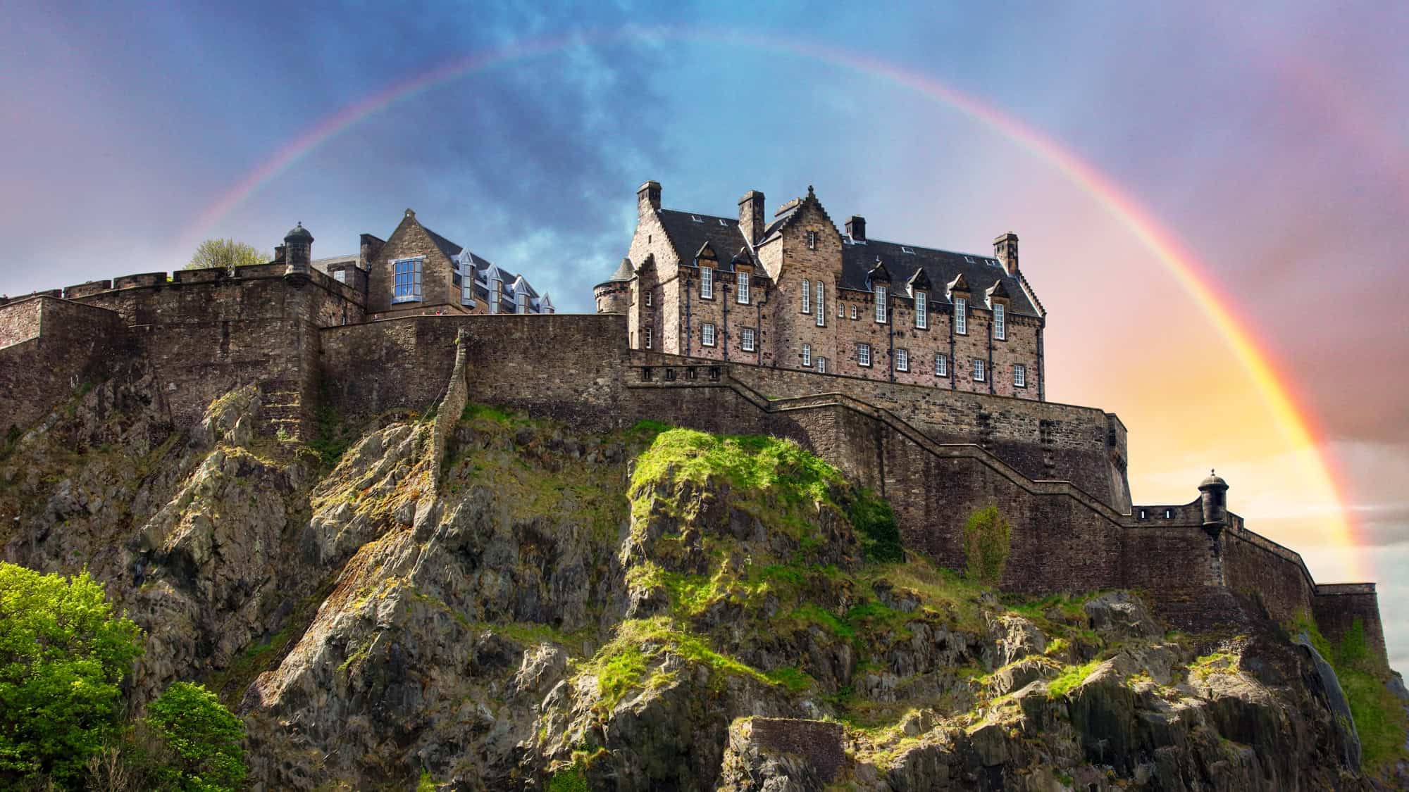Edinburgh Castle sits atop a rocky hill, highlighted by a rainbow arcing across the sky.