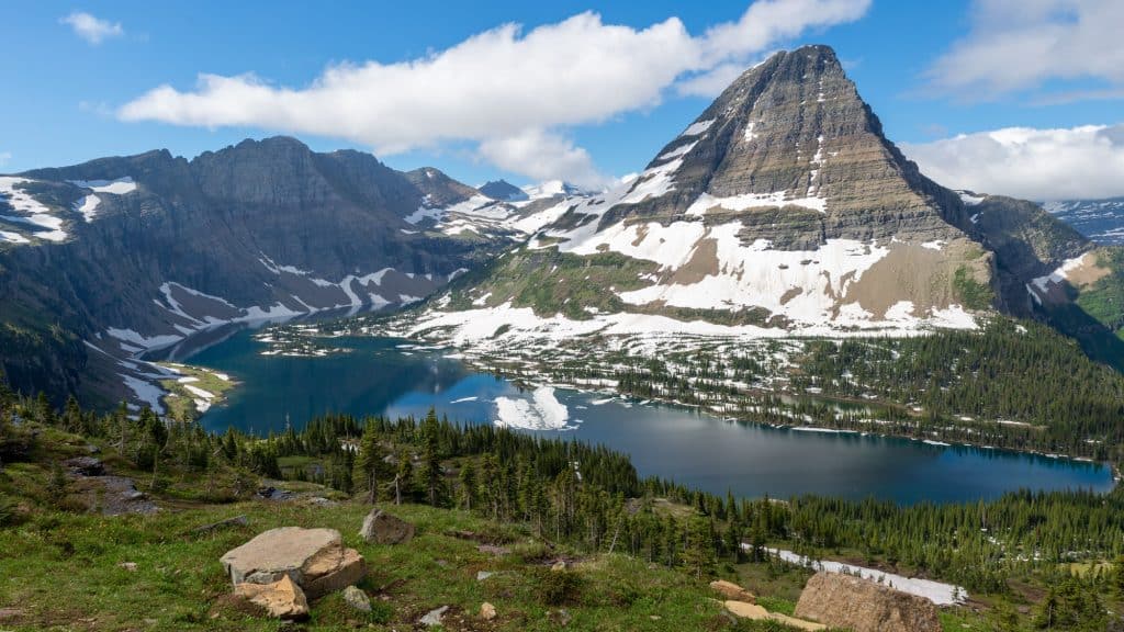 A deep blue alpine lake sits beneath snow-dusted cliffs and green ridges in a high mountain valley.
