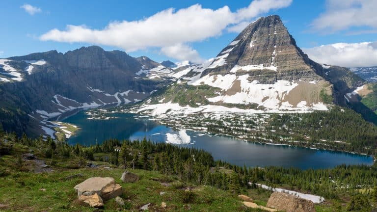 A deep blue alpine lake sits beneath snow-dusted cliffs and green ridges in a high mountain valley.