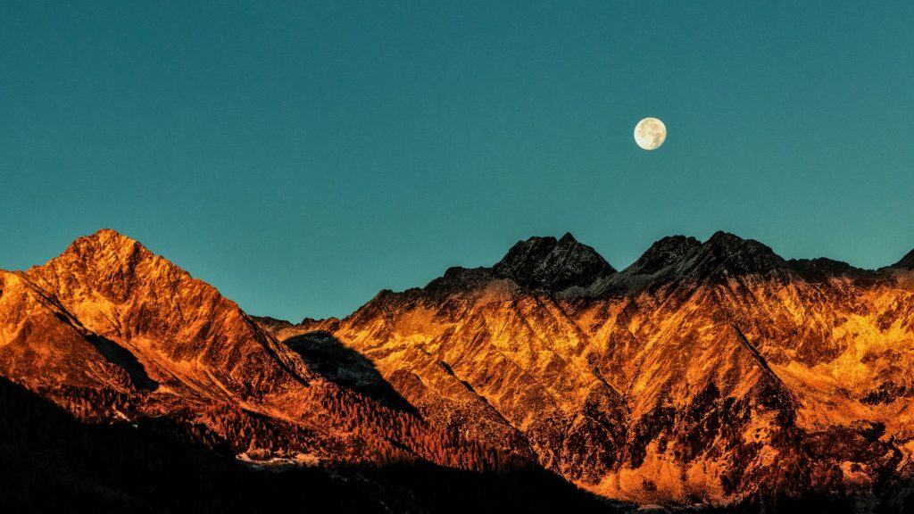 The full moon rises above jagged peaks glowing orange in the fading sunlight.