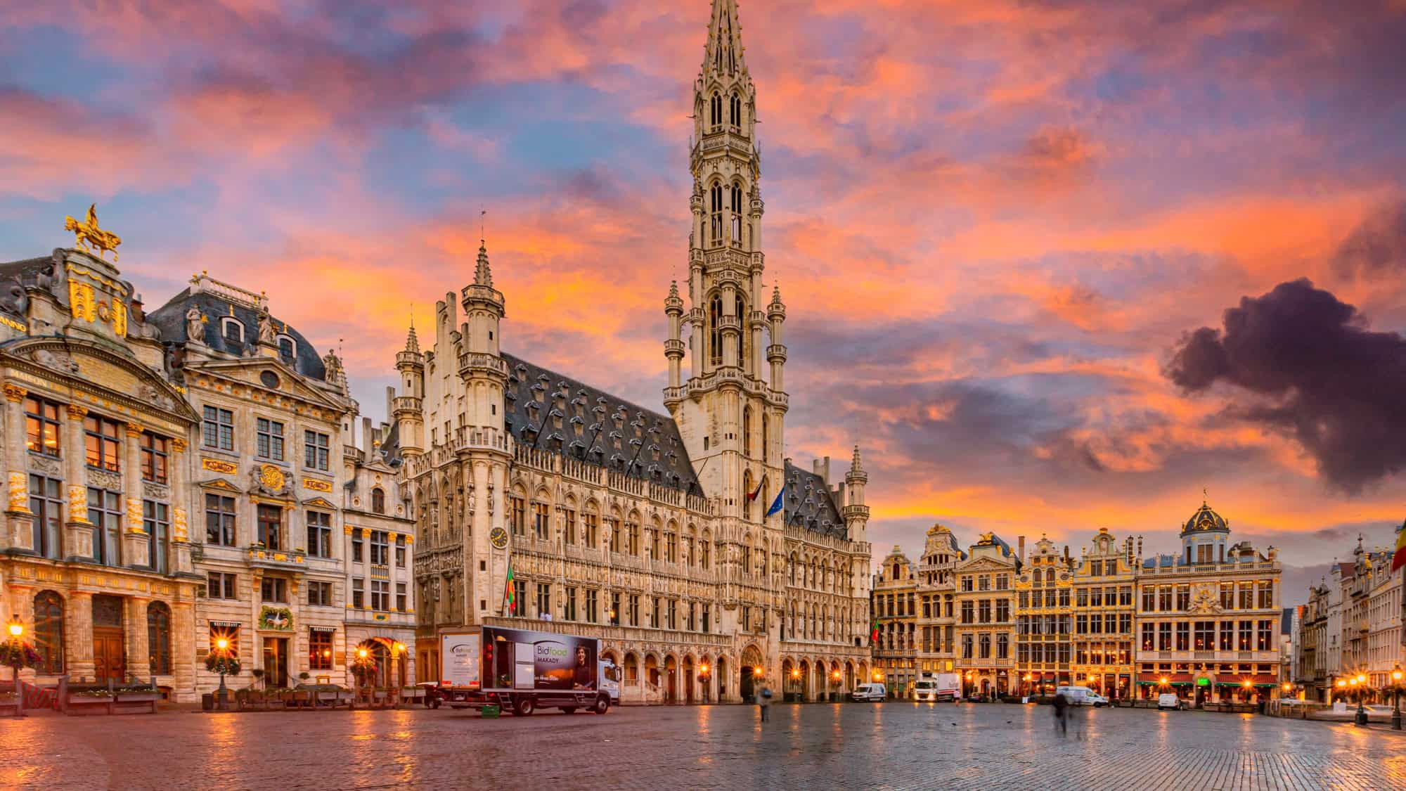The ornate Town Hall and guildhalls of Brussels’ Grand Place are illuminated beneath a dramatic sunset sky.