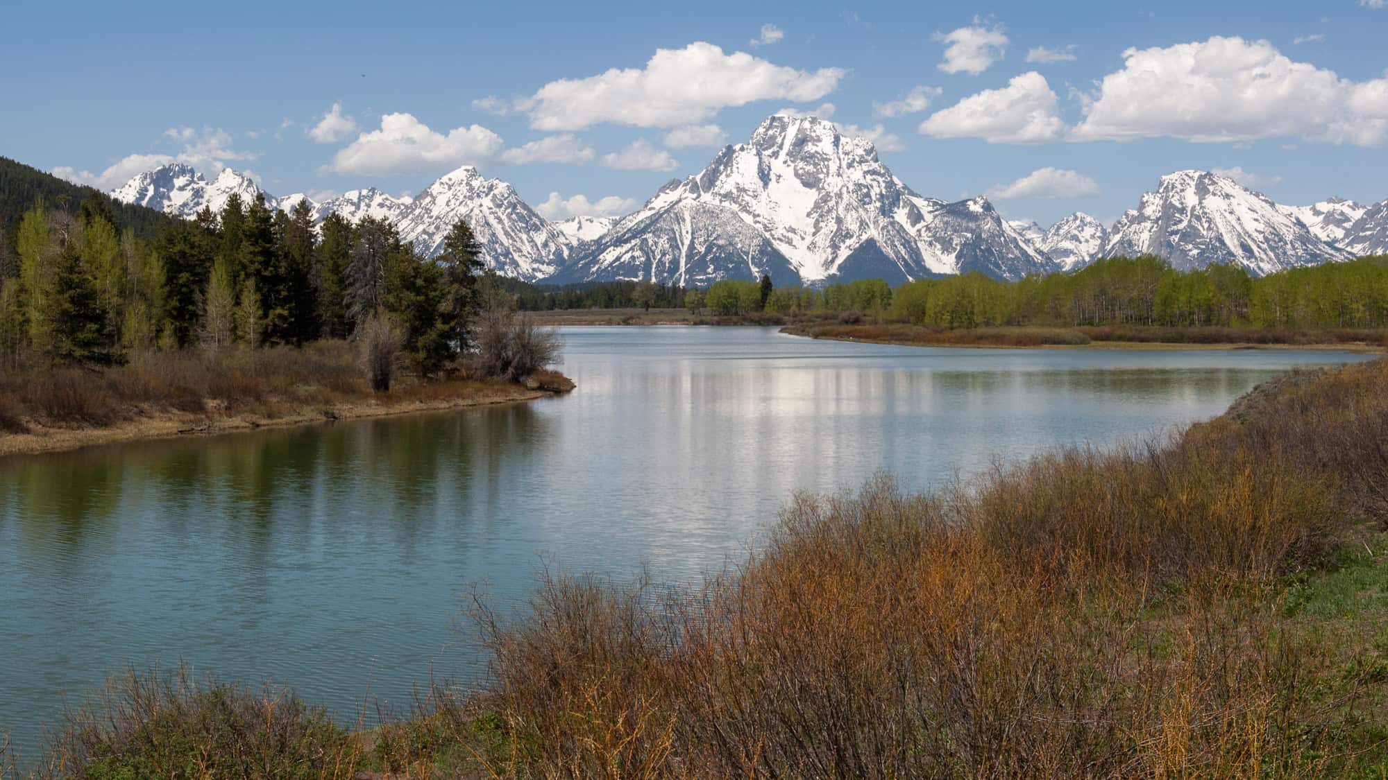A calm river winds through trees and open land with jagged, snow-covered peaks in the background.
