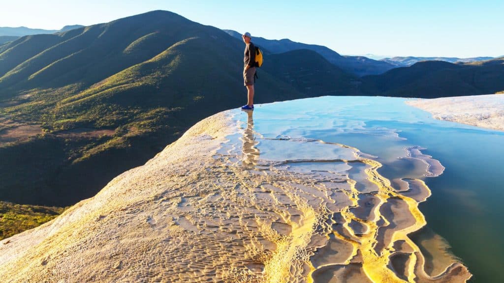 A traveler stands on the golden mineral terraces of Hierve el Agua, where reflective pools meet sweeping mountain views.