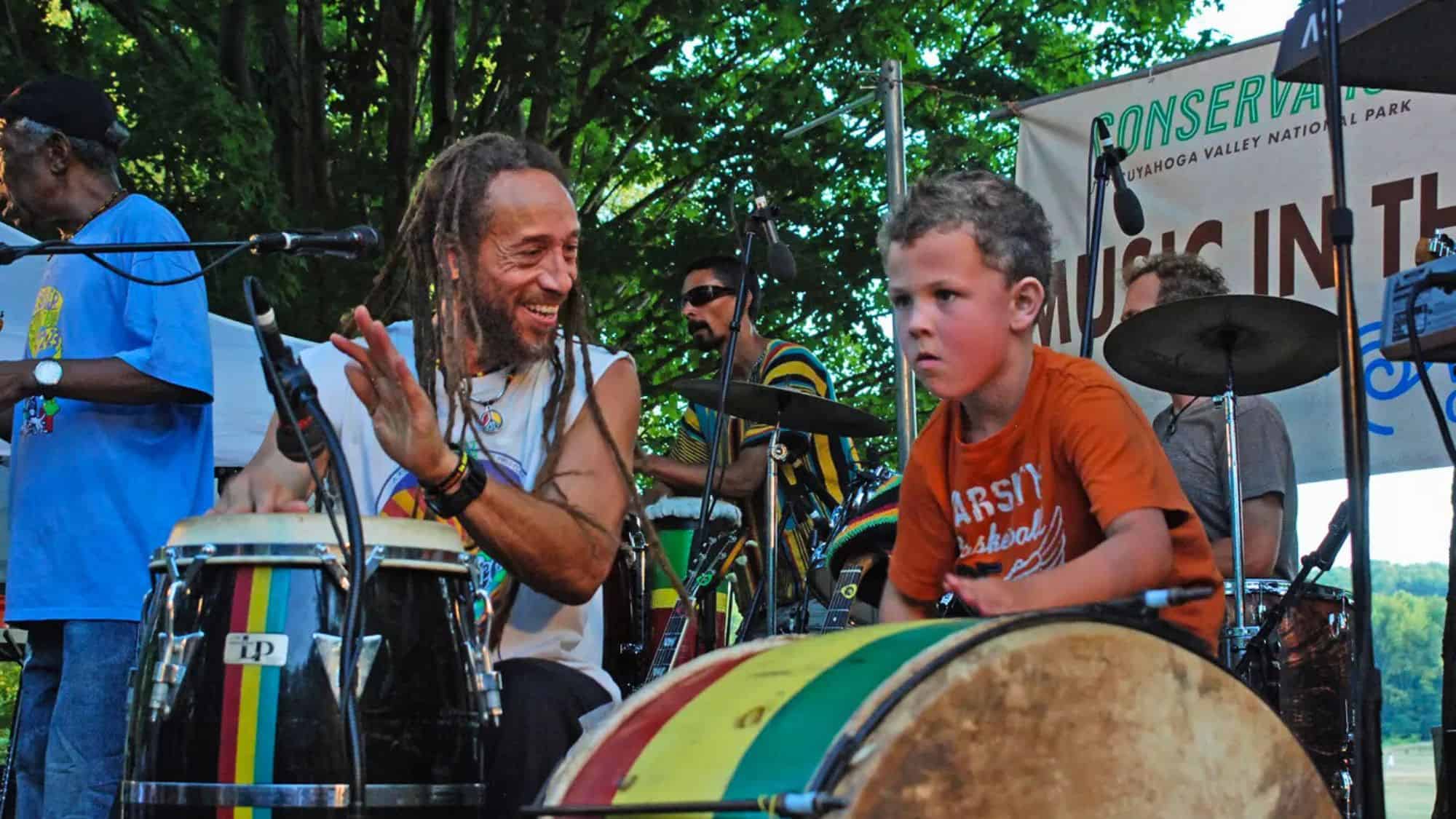 A smiling musician with dreadlocks plays conga drums alongside a focused young boy on a large drum during an outdoor music event.