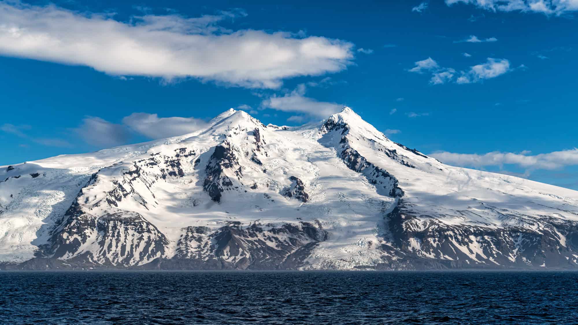 A massive volcanic peak covered in snow and ice looms above dark ocean waters under a bright sky.