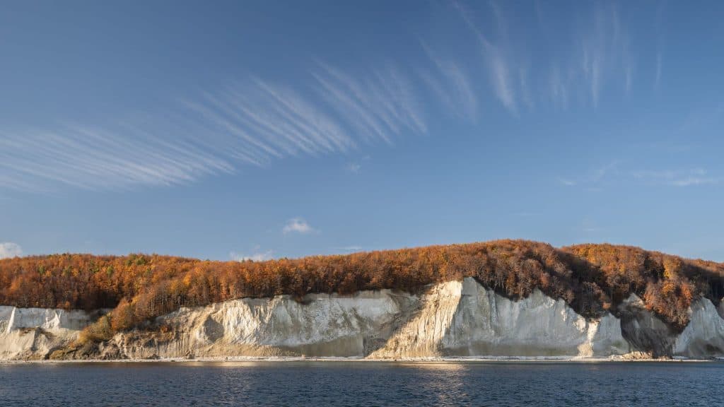 White chalk cliffs rise beneath a dense forest of autumn-colored trees, all reflected in the calm blue waters below.