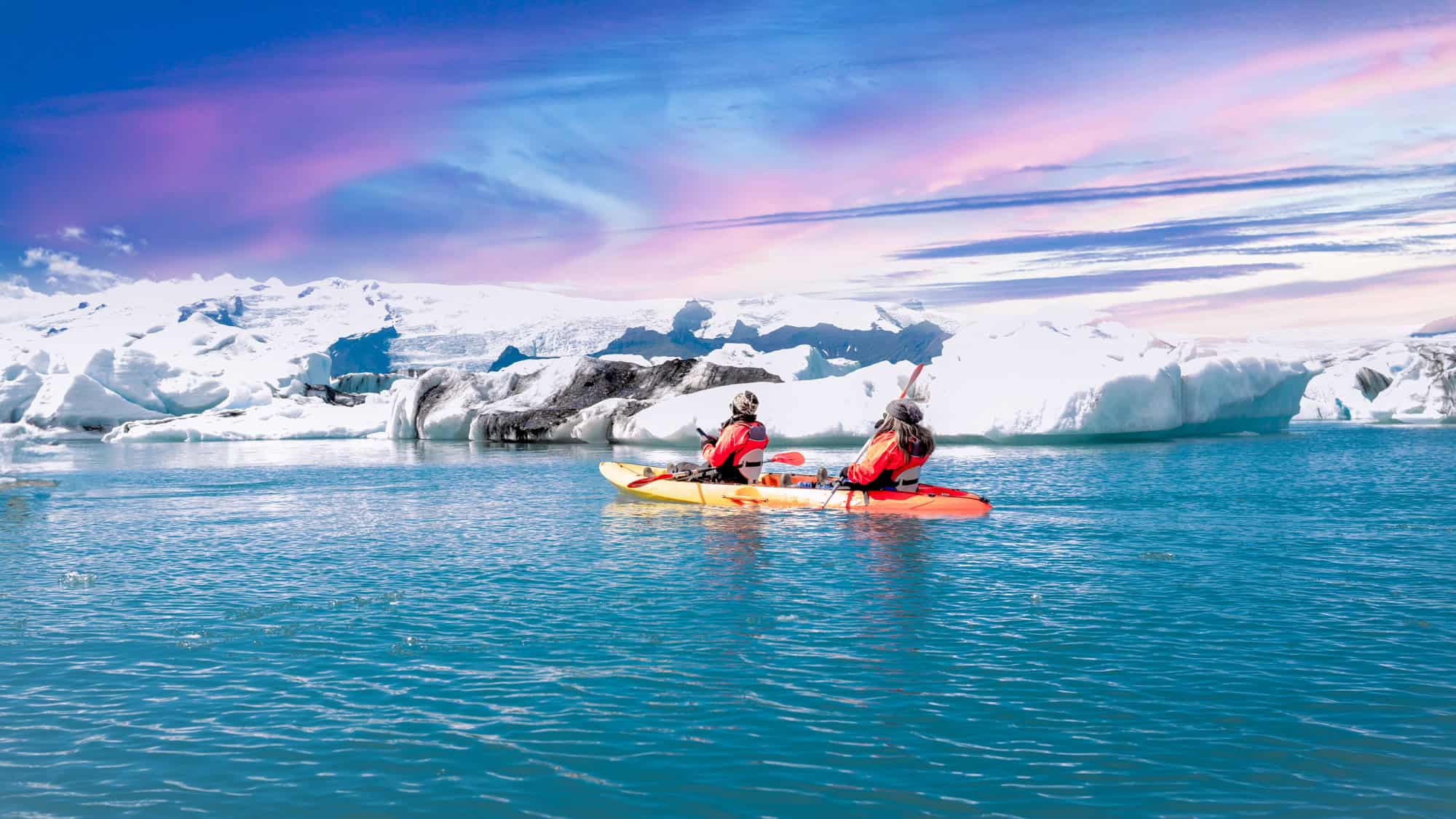 Two kayakers paddle past floating icebergs in bright orange kayaks under a vivid sky.