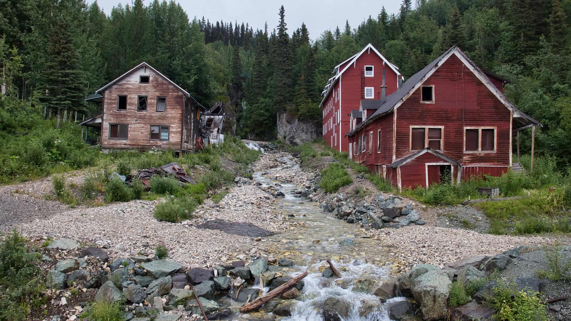 Abandoned wooden structures, including striking red mill buildings, sit beside a rocky stream in a lush, forested Alaskan valley.