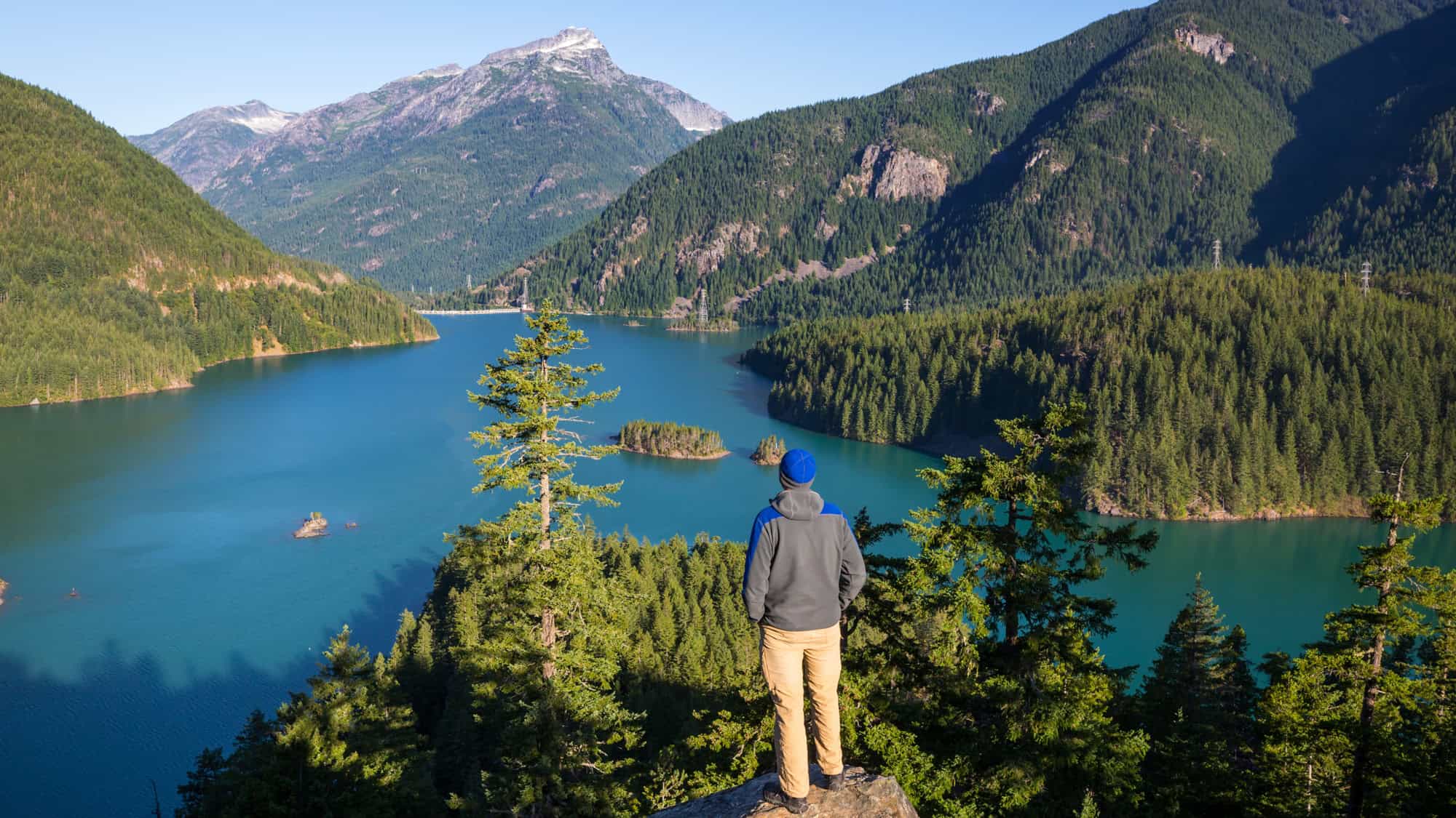 A hiker stands on a rocky ledge, gazing at a deep turquoise lake framed by steep, forested mountains.