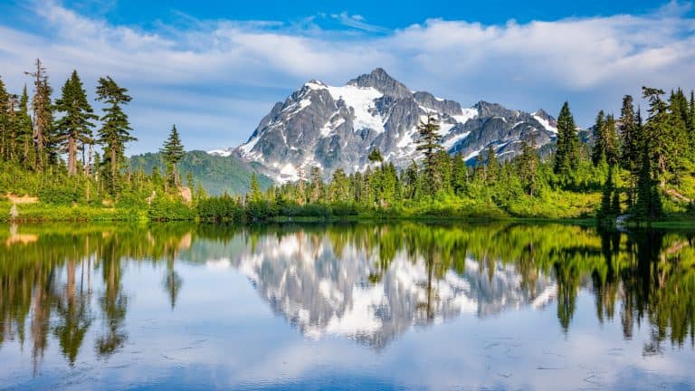 A calm alpine lake mirrors a snow-capped peak and surrounding evergreen forest under a partly cloudy sky.